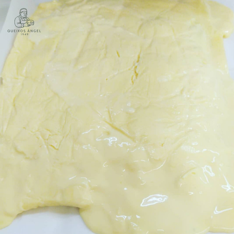 Masa de queso artesano en una superficie plana con logotipo de 'Queixos Angel' en la esquina superior izquierda. Quesos plaza de abastos Ourense. Quesería Galicia. Quesería Ourense.