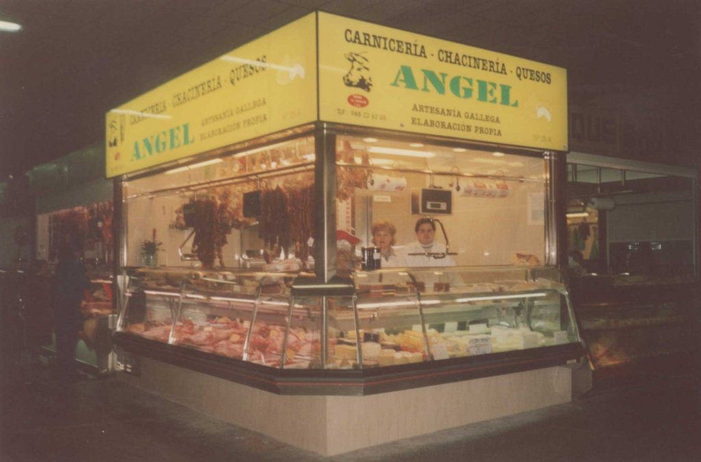 Foto antigua de puesto de venta de carnes y quesos en un mercado, con personal atendiendo y productos en exhibición, cartel que dice "Carnicería, Chacinería, Queixos, Angel".