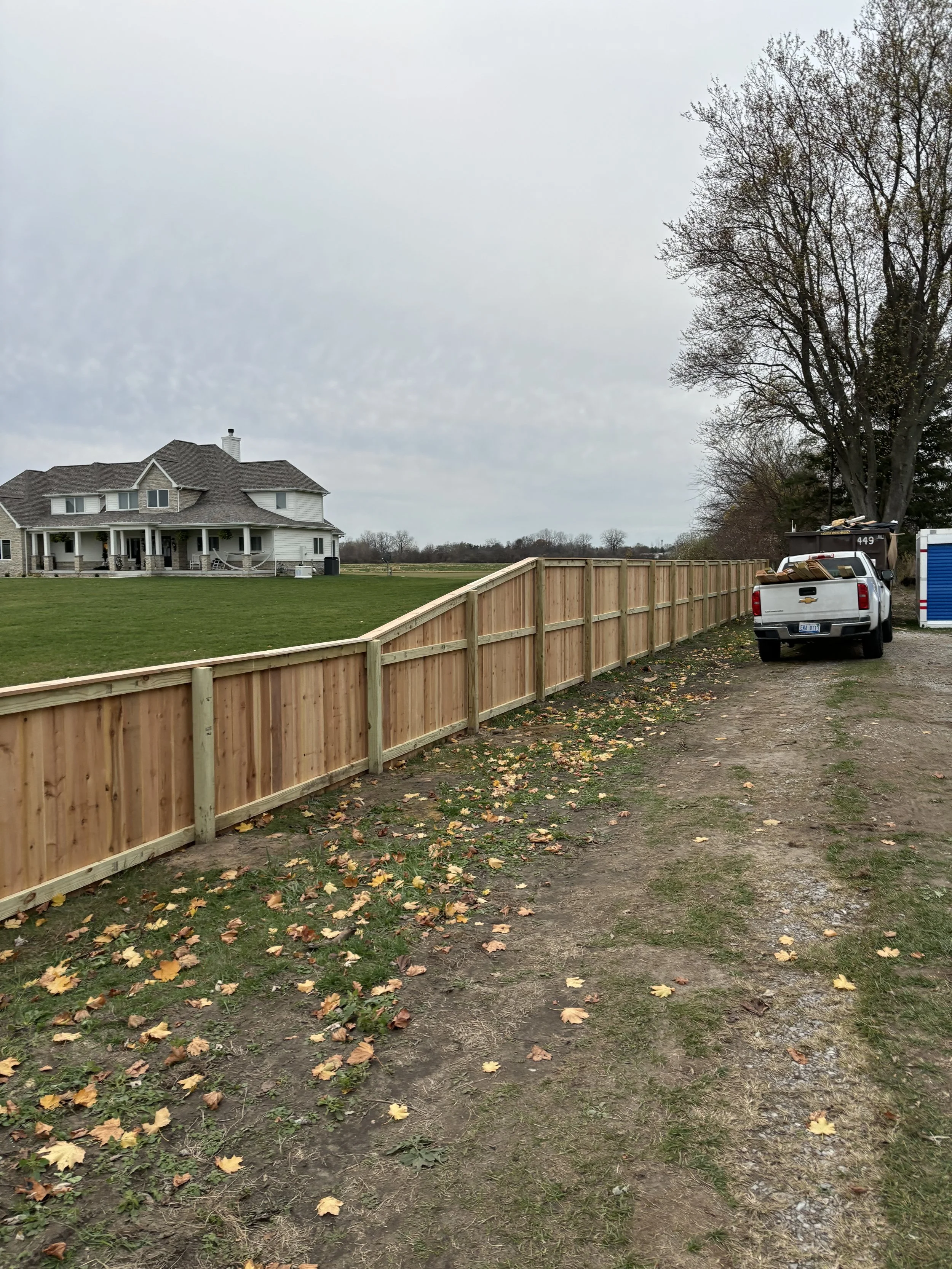 Landscaping in progress with a newly installed wooden fence along a dirt path near a house and trees, on a cloudy day.