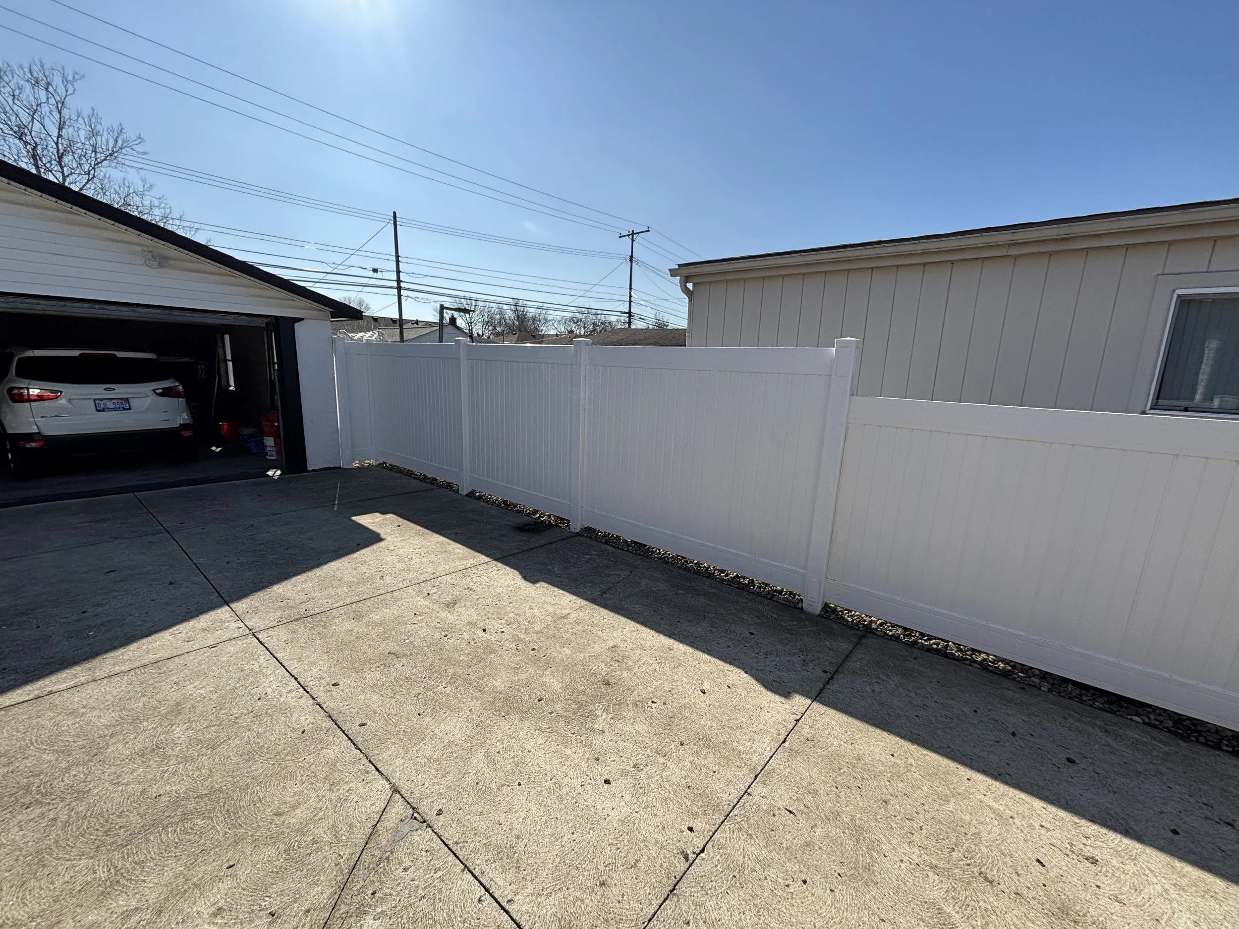 A paved backyard with a white vinyl fence and a garage with a silver car parked inside, in a suburban neighborhood with visible power lines and a clear blue sky.
