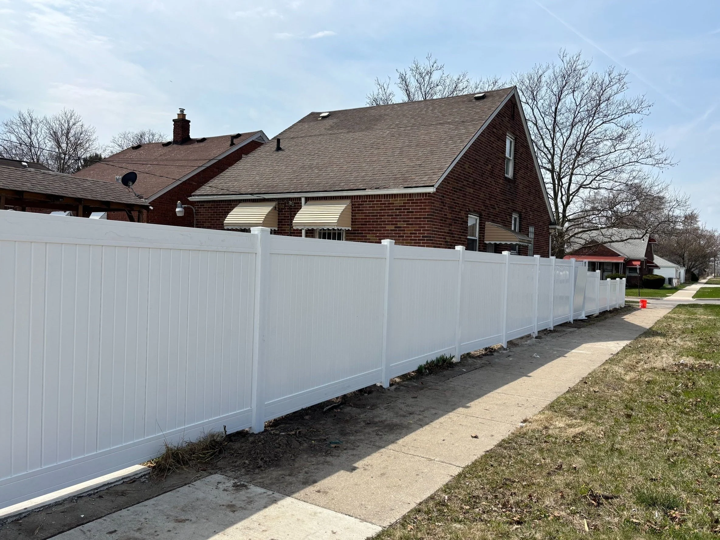A white vinyl fence running along the sidewalk outside a red brick house with a gray shingle roof and awnings over the windows, on a sunny day with some clouds and leafless trees in the background.