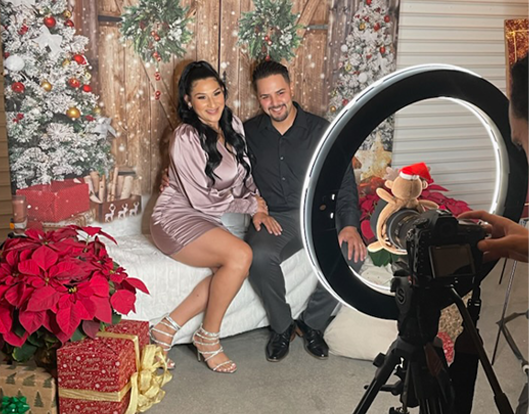 Couple posing for a holiday portrait on a white couch in front of Christmas trees and poinsettias while a photographer captures them with a ring light and camera. OC Videography holiday, photobooth, and portrait photo services.