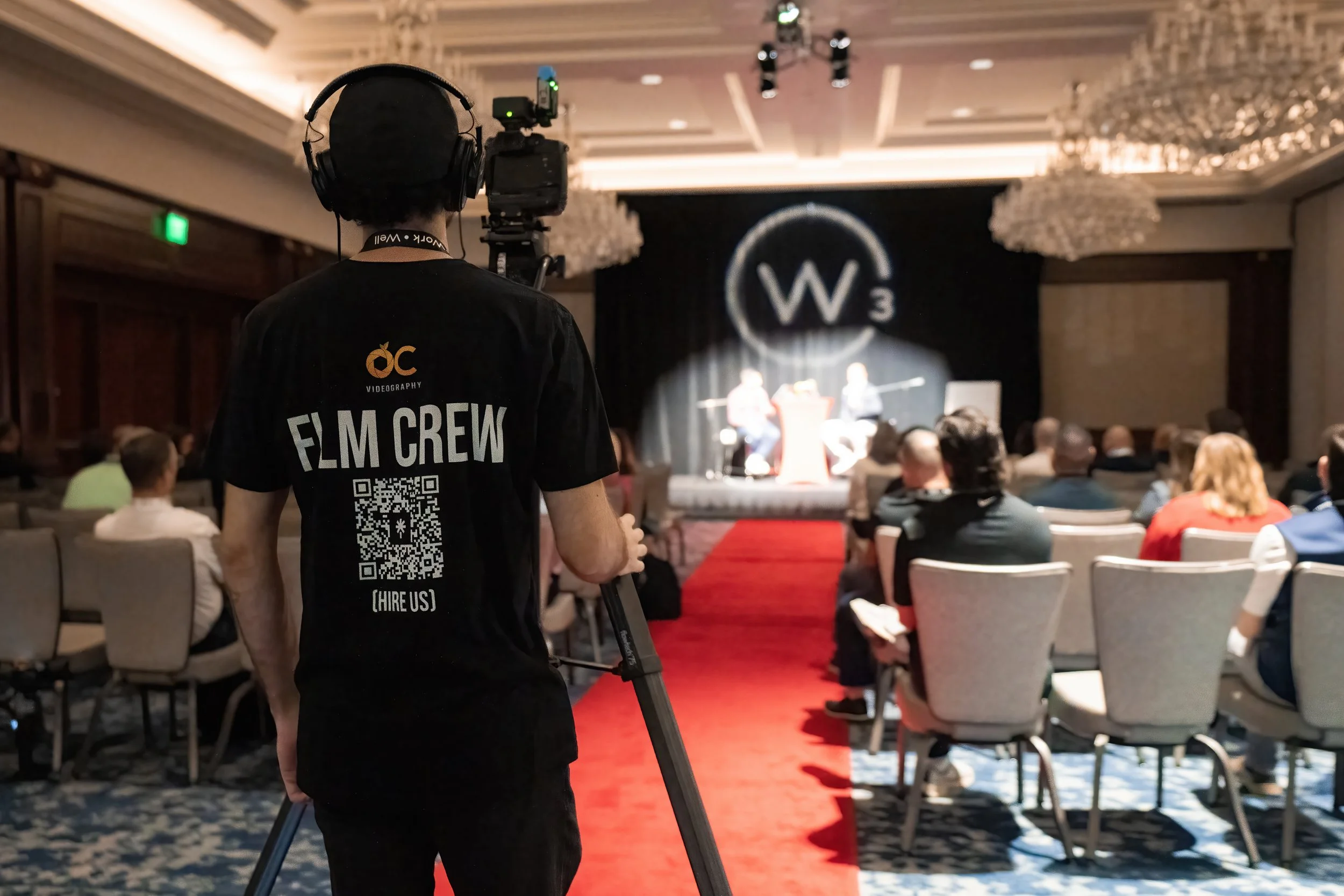 Videographer in black FILM CREW shirt filming a live event panel discussion in a conference hall with chandeliers. Corporate event video coverage by OC Videography.