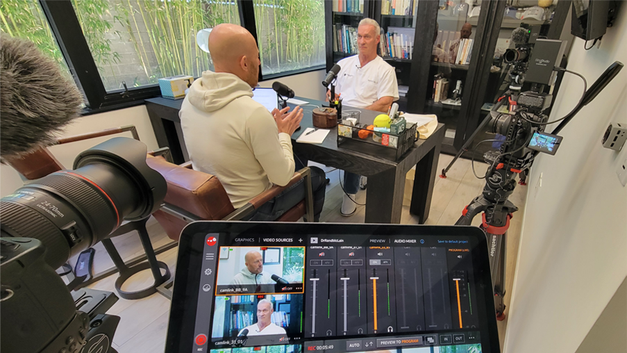 Professional interview filmed by OC Videography featuring a man in white shirt speaking on camera amid lighting, microphones, and bookshelves in a studio environment.