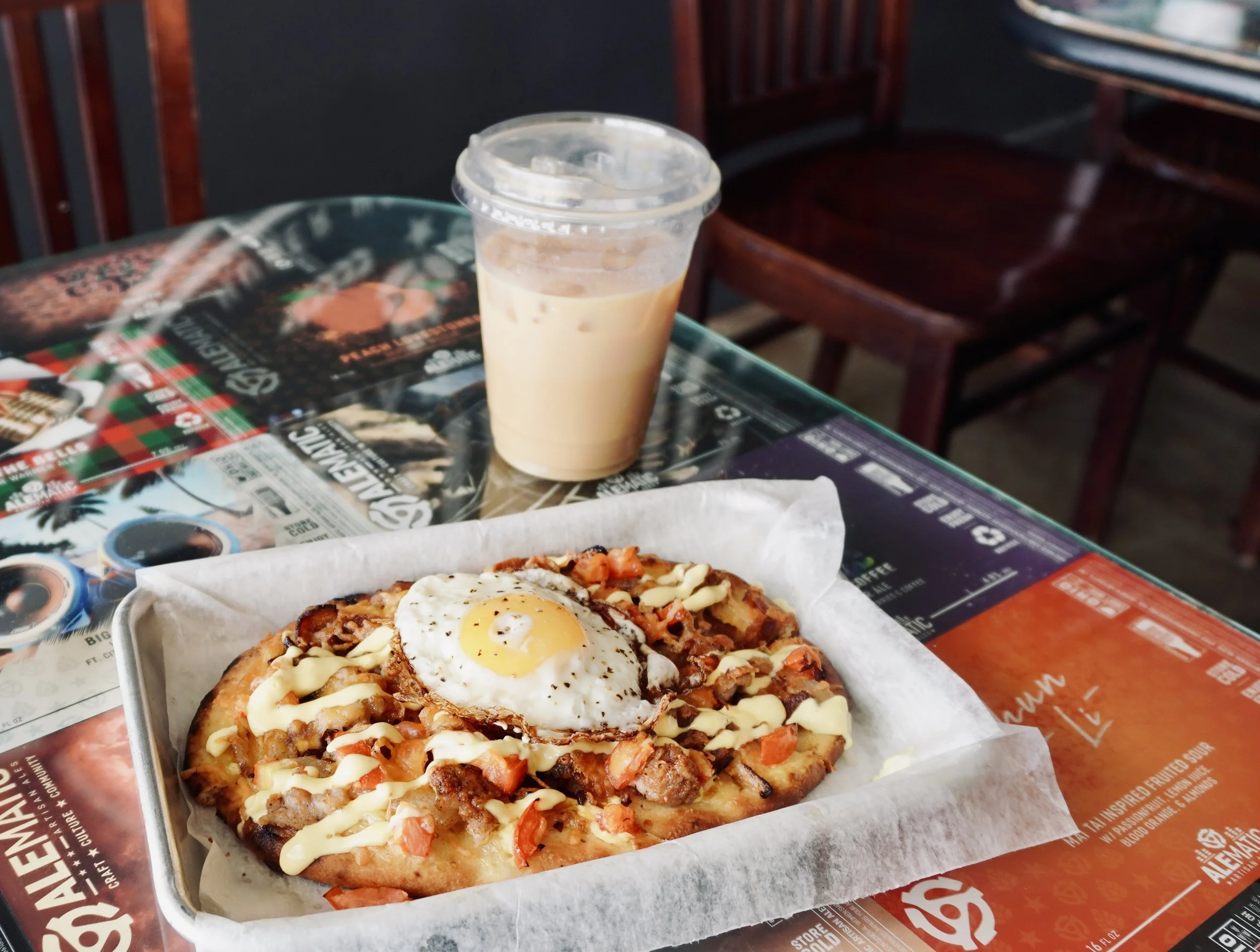 A pizza topped with an egg and various ingredients, served in a tray lined with parchment paper, alongside a plastic cup of iced coffee on a colorful table at a restaurant.