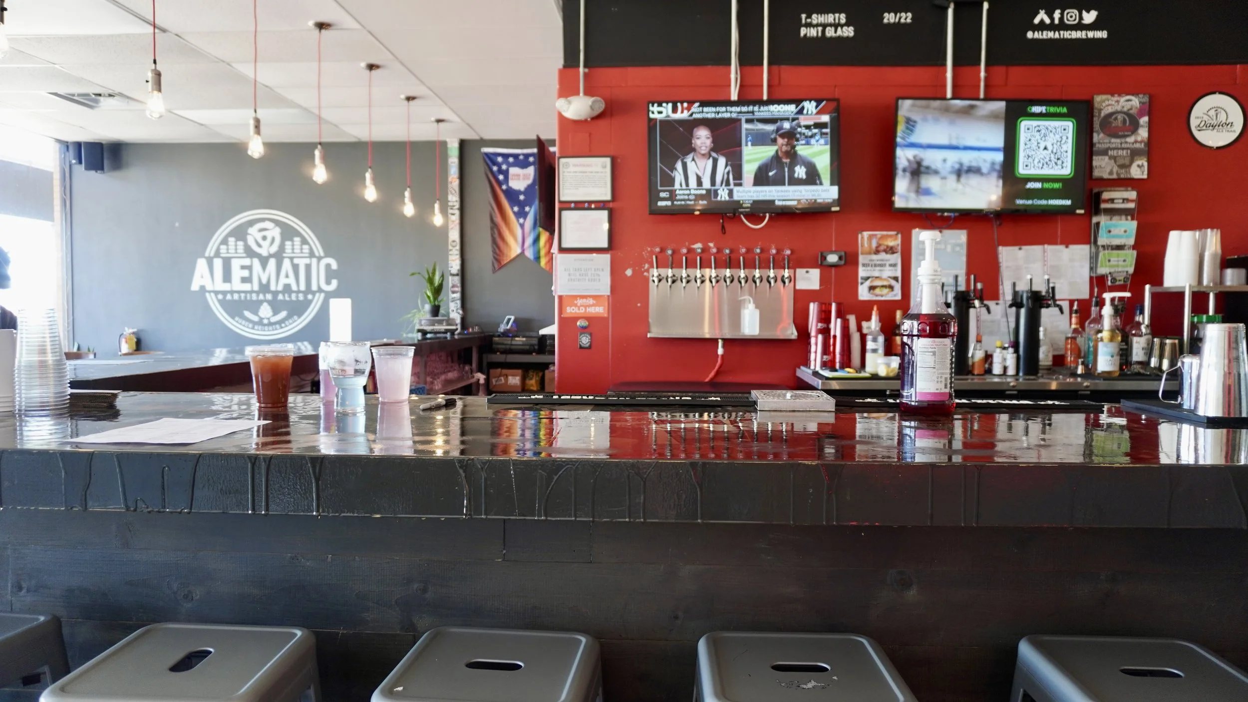 Interior of a bar or brewery with a polished wooden bar counter, drinks, and bar equipment. There are multiple TV screens on a red wall, one displaying sports and another a QR code. A large Alematic Brewing logo is on a gray wall in the background.