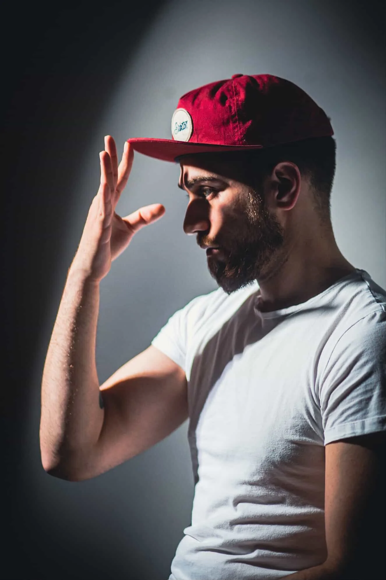 A man with a beard wearing a white t-shirt and a red baseball cap, looking down with his hand adjusting the cap.
