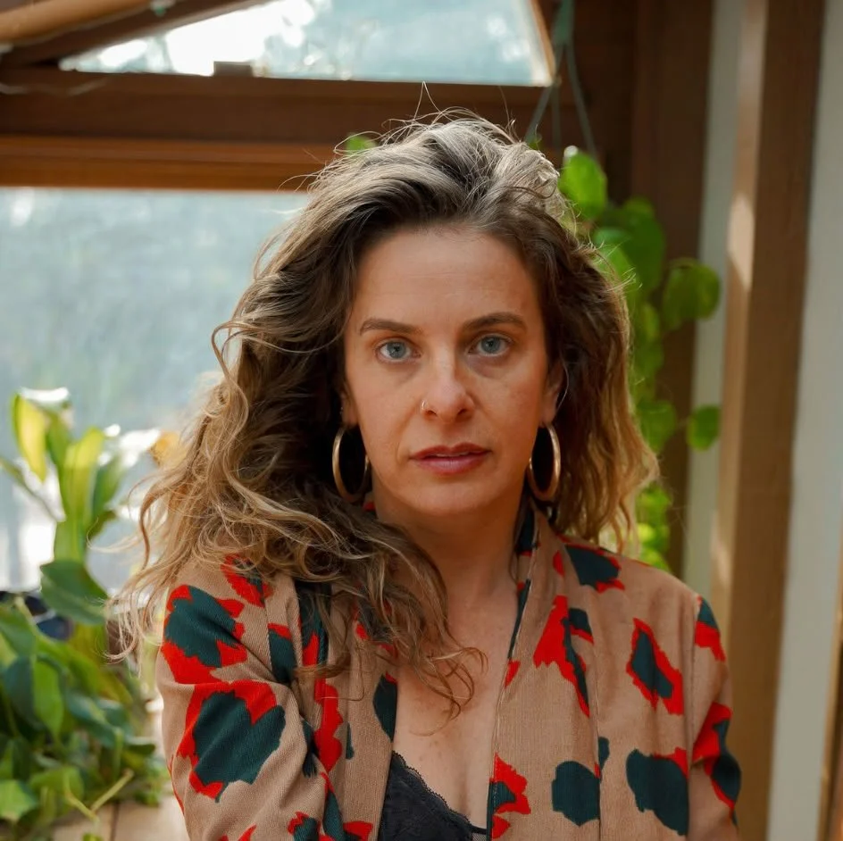 A woman with wavy brown hair and blue eyes, wearing large hoop earrings and a leopard-print blazer, sitting indoors near a window with plants visible in the background.