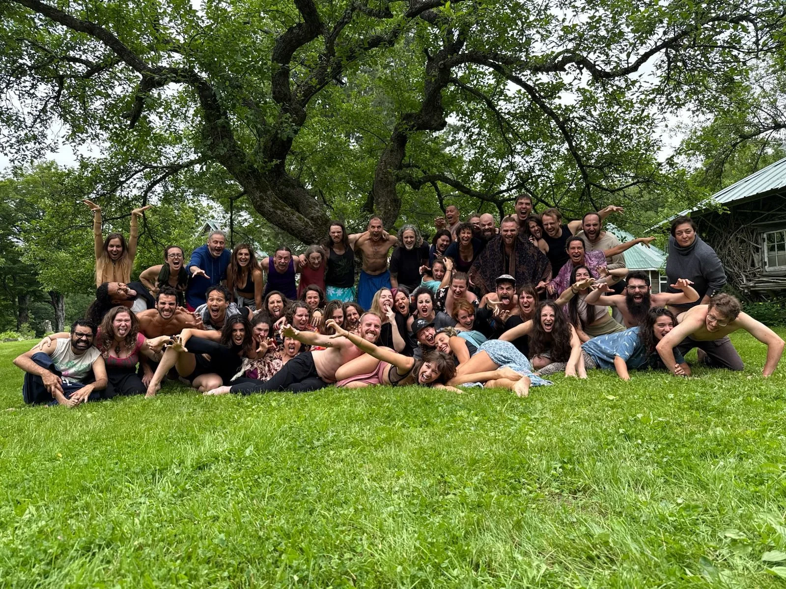 Group of people posing and laughing outdoors on a grassy field with trees and a shed in the background.