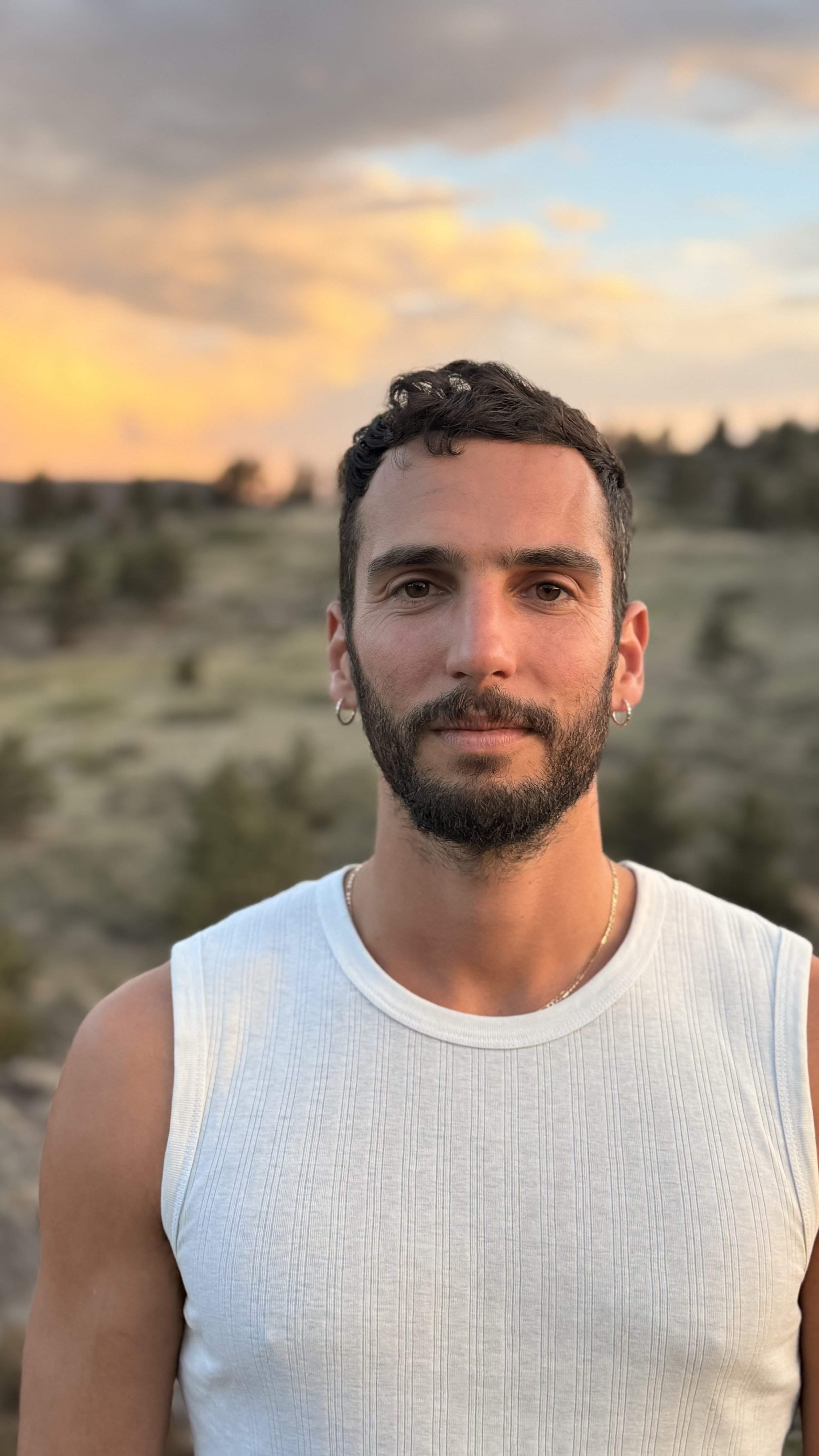 A man with a shaved head and stubble beard takes a selfie outdoors, with rocky terrain and green leaves in the background.