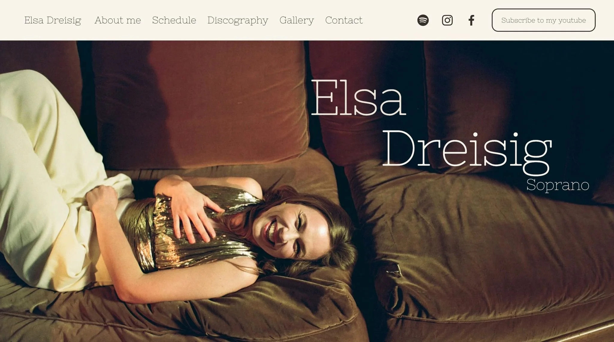 A woman lying on a brown couch, smiling and wearing a shiny, metallic gold top and cream-colored pants.