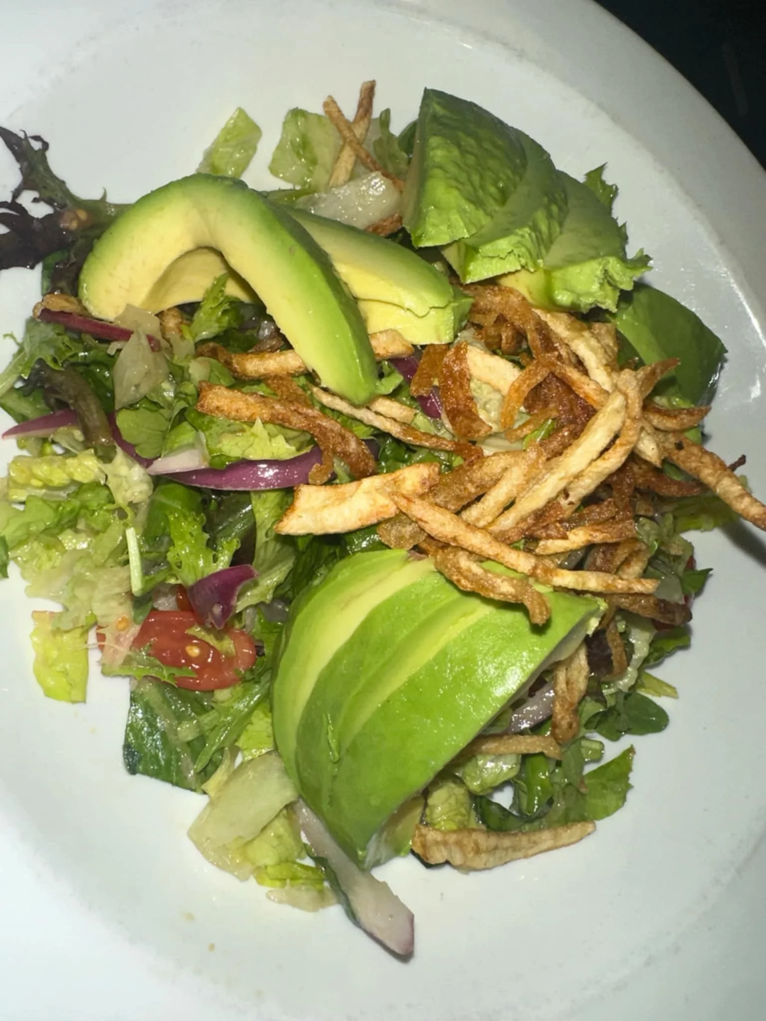 Mixed salad with sliced avocado, crispy fried strips, lettuce, tomato, cucumber, and onions on a white plate.