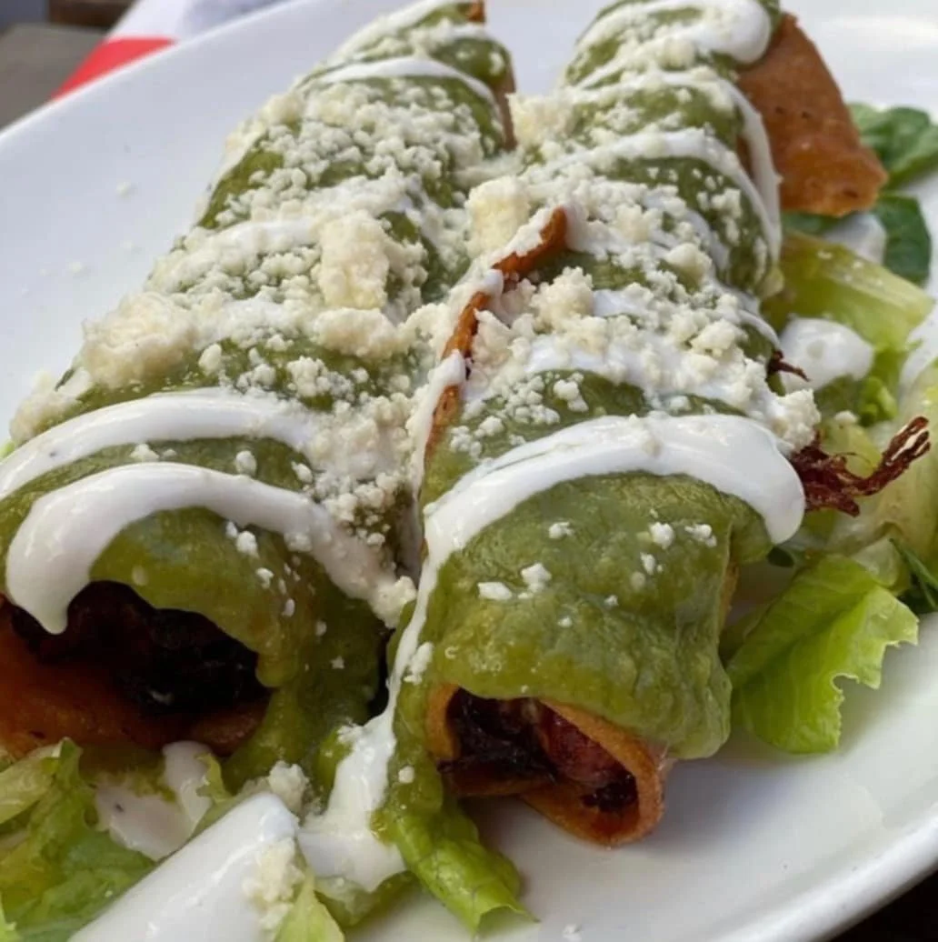 Close-up of a plate of Mexican taquitos topped with sour cream, shredded cheese, and guacamole, served on a bed of lettuce.