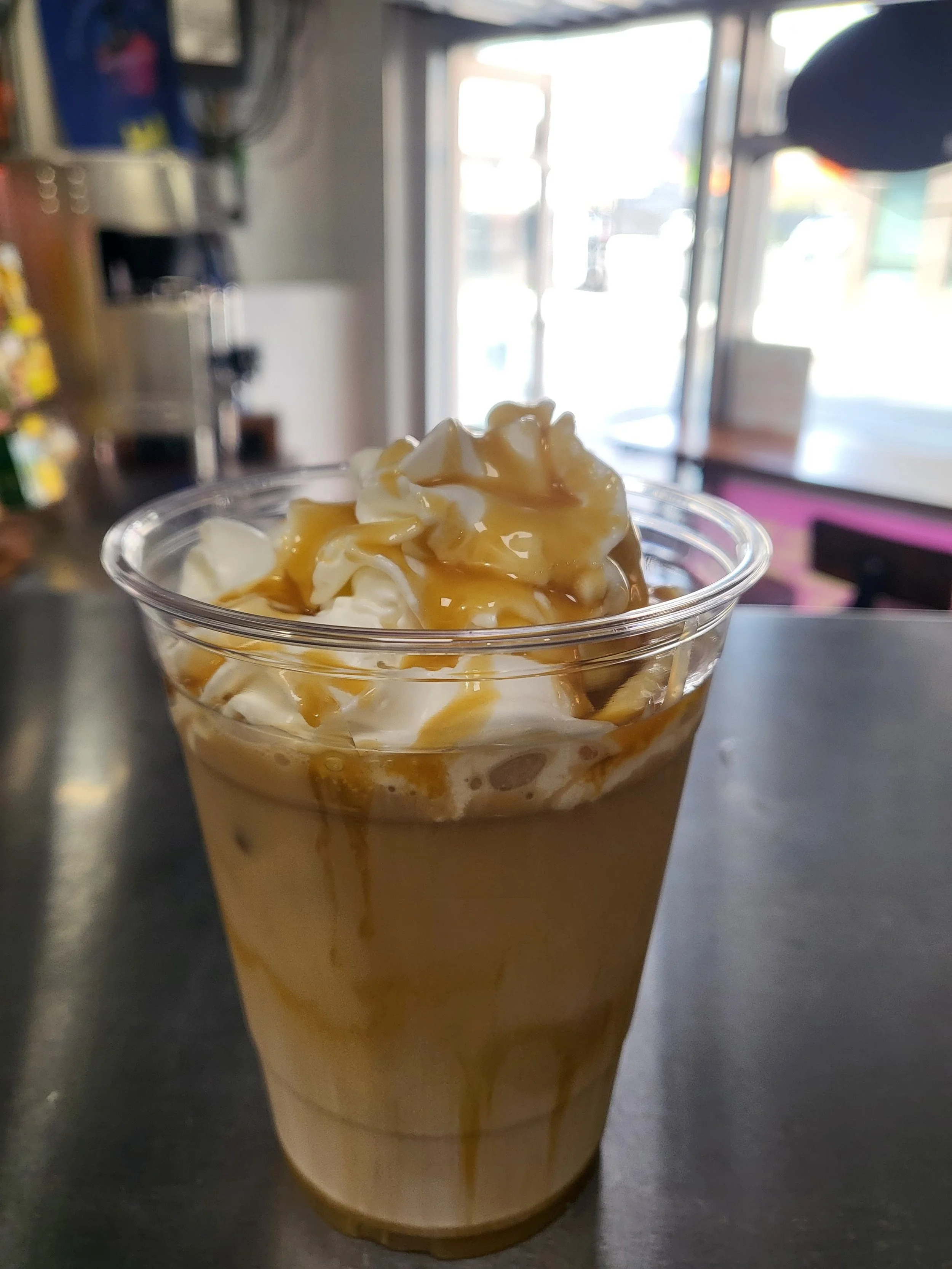 Caramel iced coffee topped with whipped cream and caramel drizzle inside a clear plastic cup, on a dark countertop inside a café.