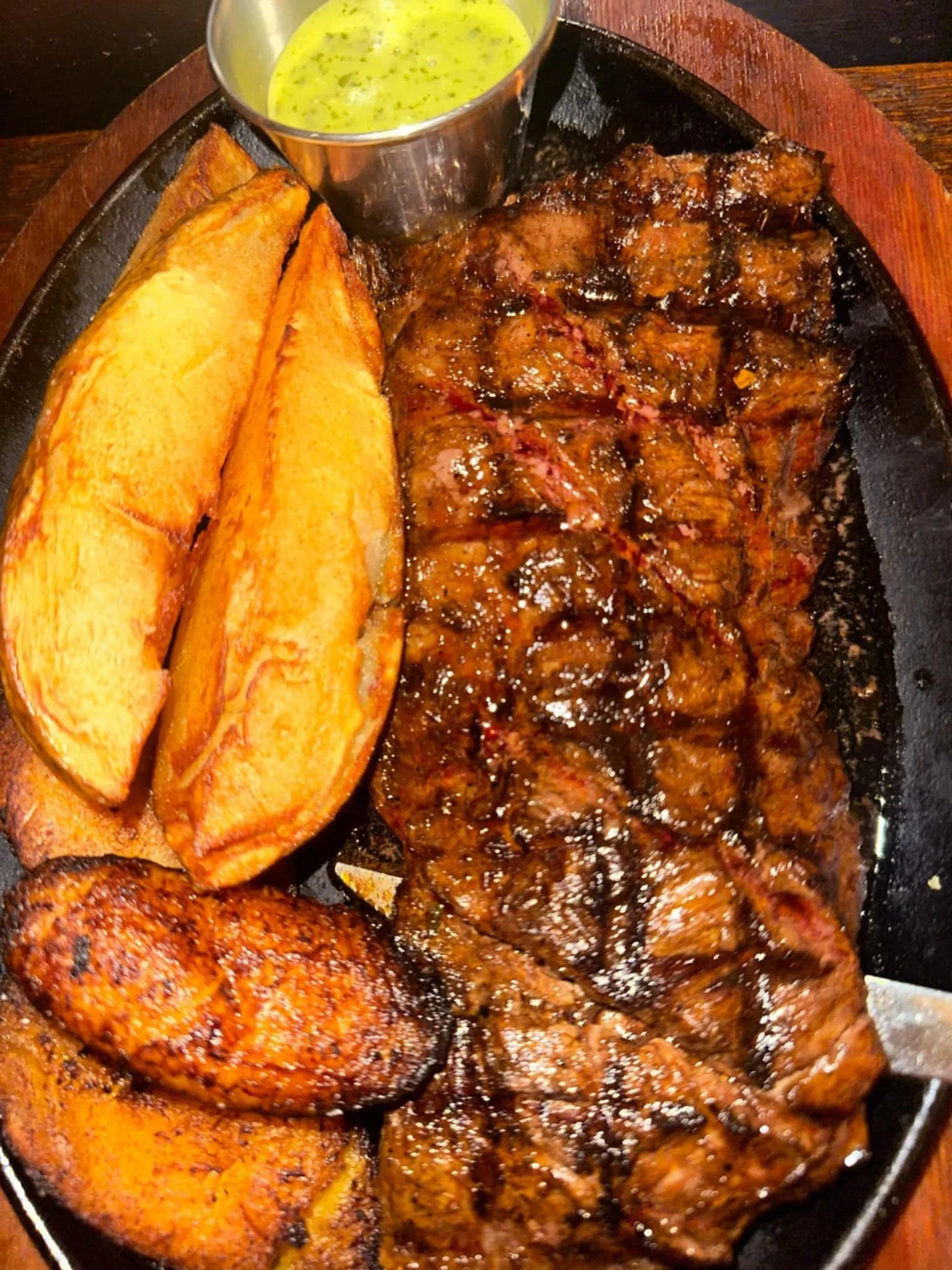 A sizzling steak topped with sauce, two large potato wedges, a fried chicken wing, and a small cup of green sauce on a hot cast iron skillet.