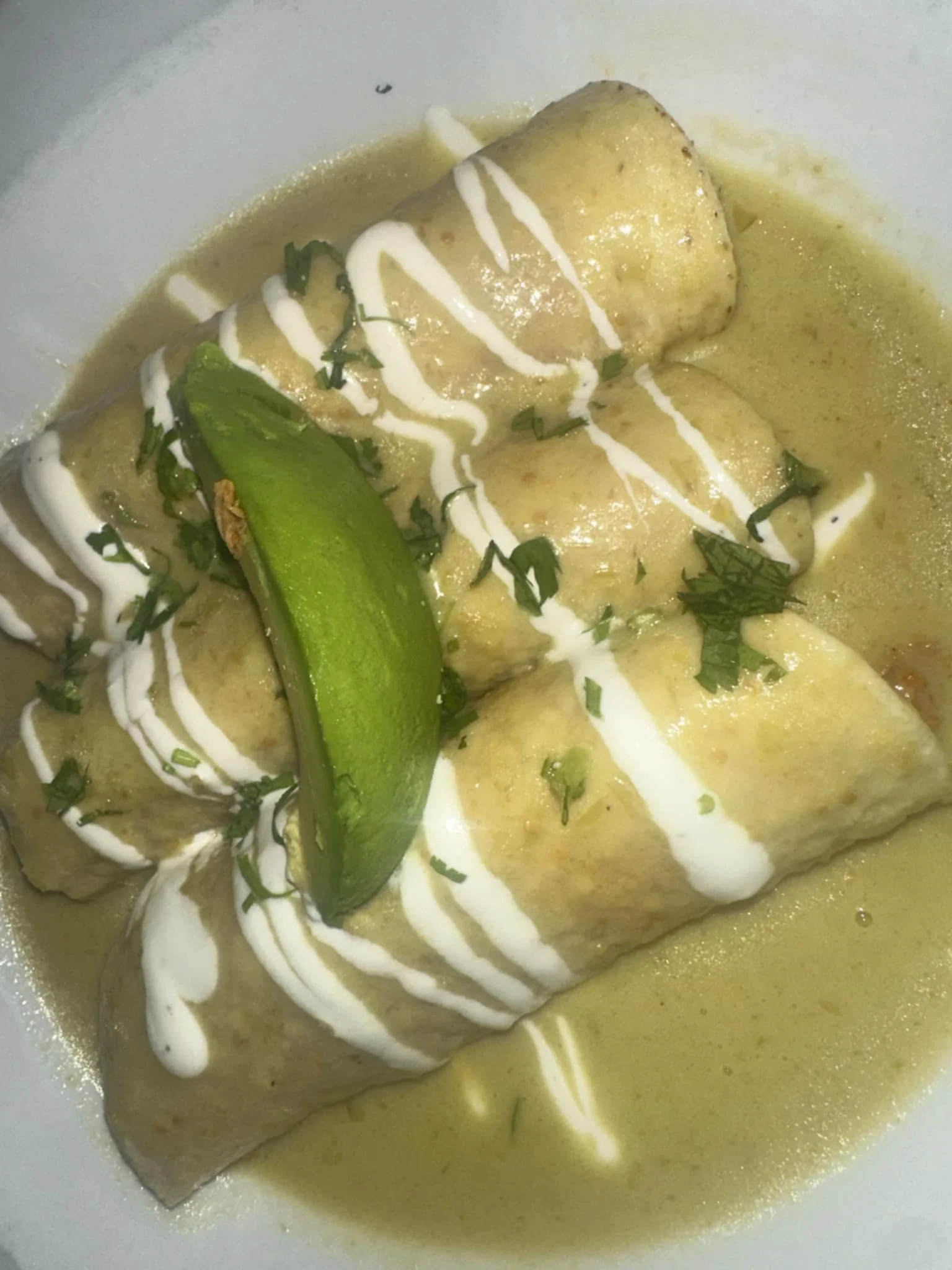 Close-up of three stuffed enchiladas topped with green chili sauce, white crema drizzle, and chopped herbs, garnished with a lime wedge.