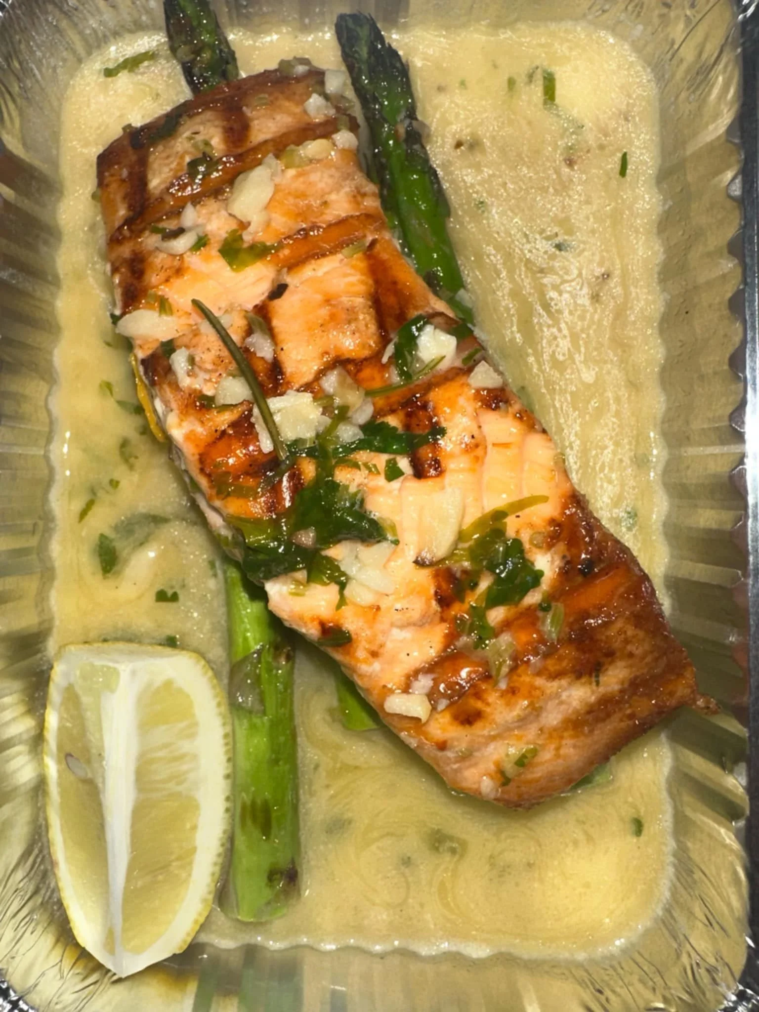 Baked salmon fillet topped with herbs and onions, served with asparagus and a lemon wedge in a creamy sauce.
