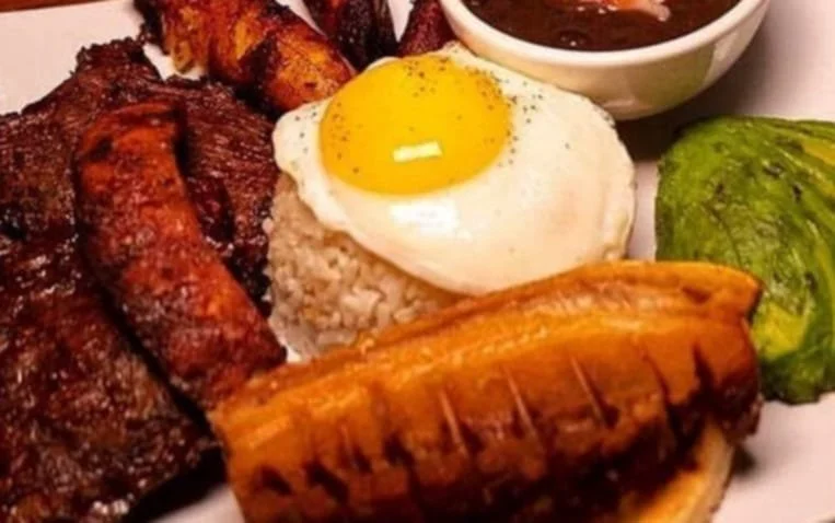 Plate of grilled meat, rice with a fried egg on top, fried plantain, roasted banana, avocado, and a side of beans.