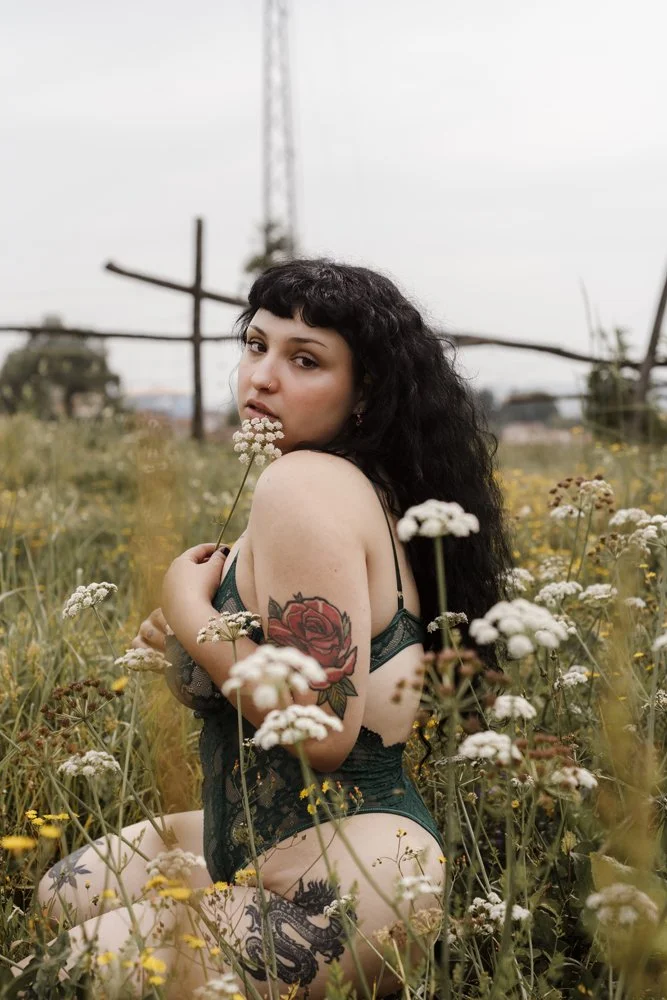 woman standing in a field, wearing white lingerie and a thin black leather harness on her torso.