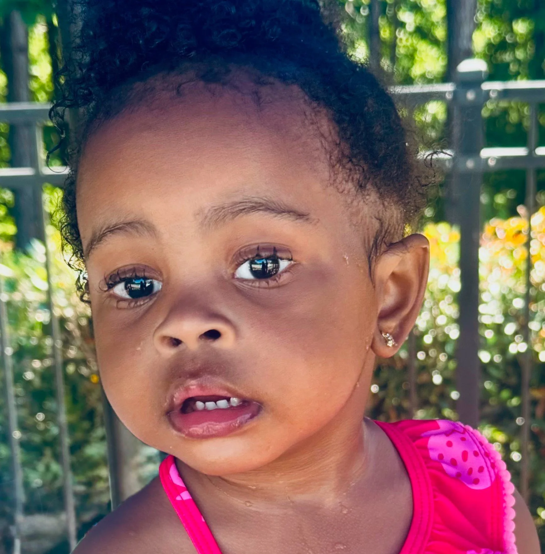 Close-up of a young girl with curly hair, wearing a pink swimsuit with dotted heart design, standing outdoors near a black metal fence, with greenery and sunlight in the background.
