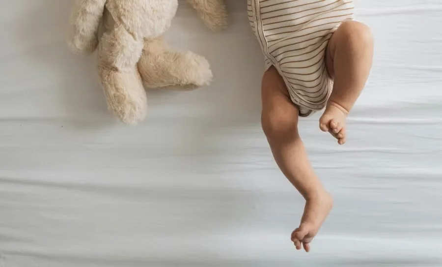 A baby in a striped onesie lying on a white bed next to a cream-colored teddy bear.