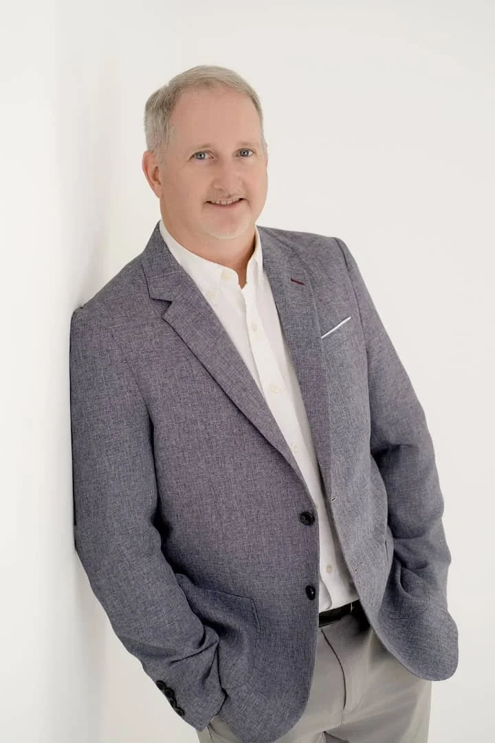A middle-aged man with short gray hair and a beard, wearing a gray blazer over a white shirt, standing against a white wall, smiling at the camera.