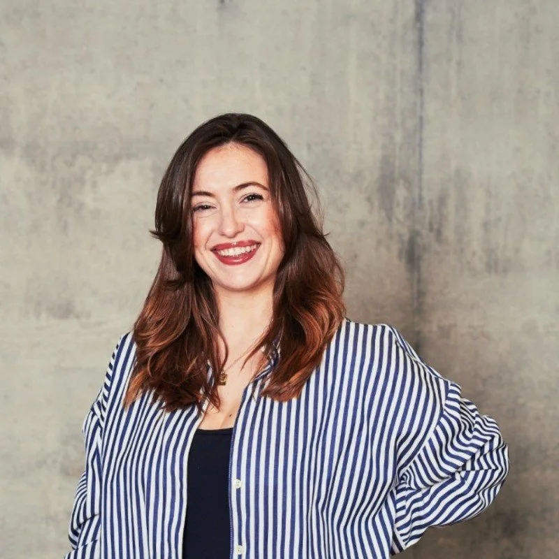A woman with long dark hair, wearing a black top with pink floral patterns, standing against a neutral background for Molly Muratov's business coaching website. 