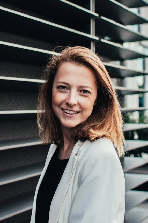 A woman with shoulder-length blonde hair smiling, wearing a black blazer over a brown top, standing against a textured blue-green background for Molly Muratov's business coaching website. 