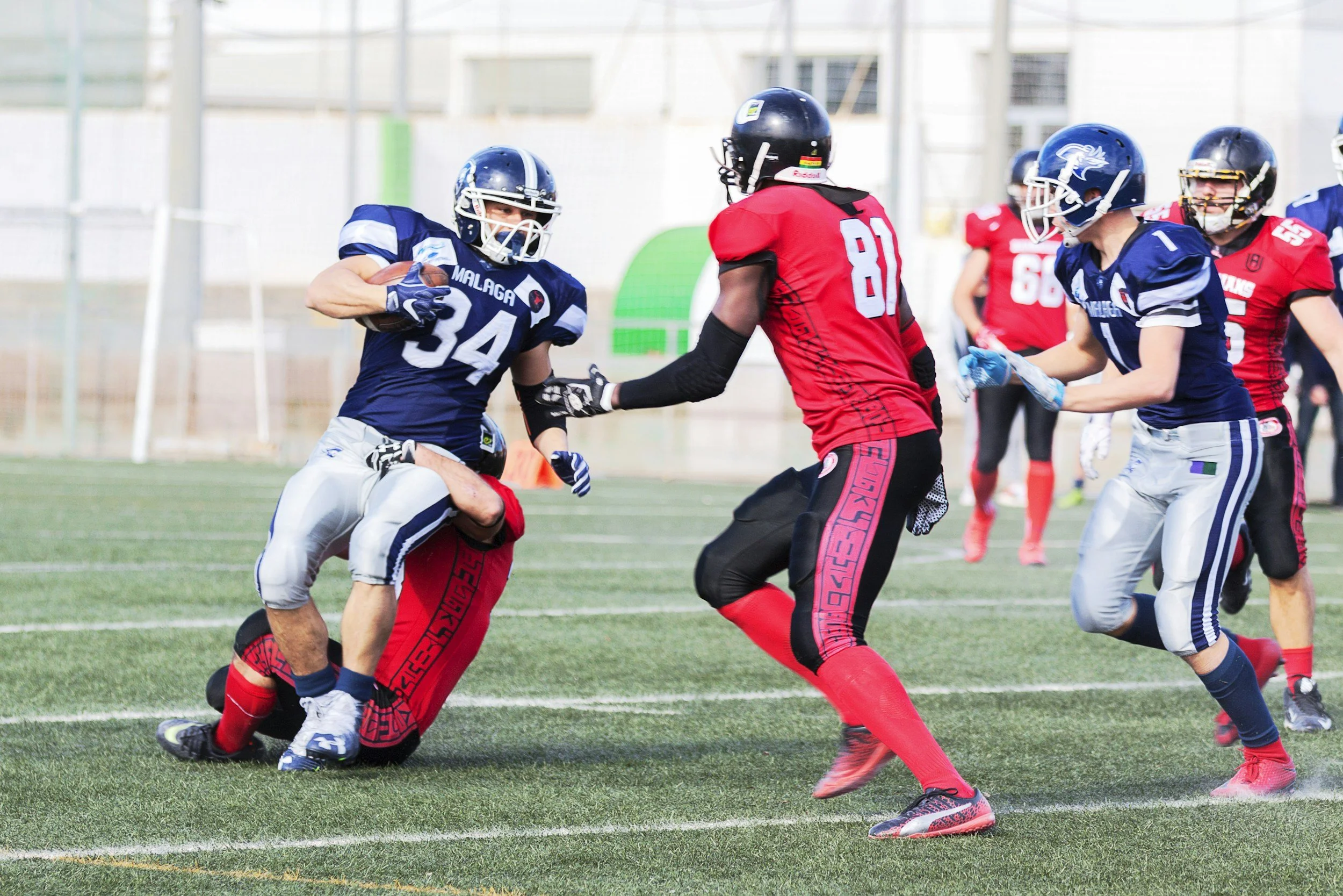 American football game with players in blue and red jerseys. One player in blue is running with the ball, while another blue player and a red player engage in blocking.