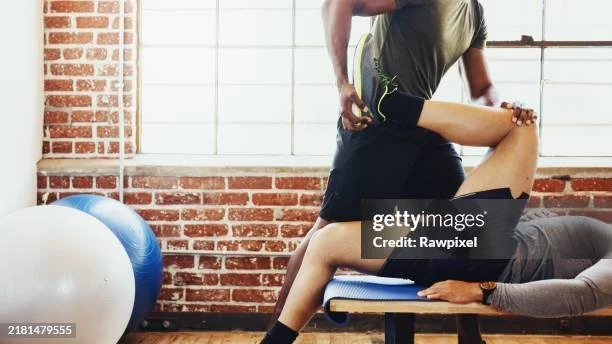 A physical therapist providing leg stretching exercise to a patient lying on a treatment table in a gym or clinic with brick walls.