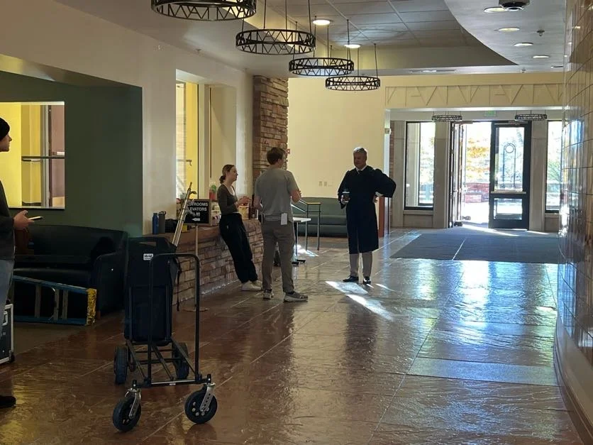 Interior of a building lobby with three people talking near the entrance, a man in a suit, a woman, and a woman with a ponytail, with a small table and some luggage in the background, and sunlight coming through the glass doors.