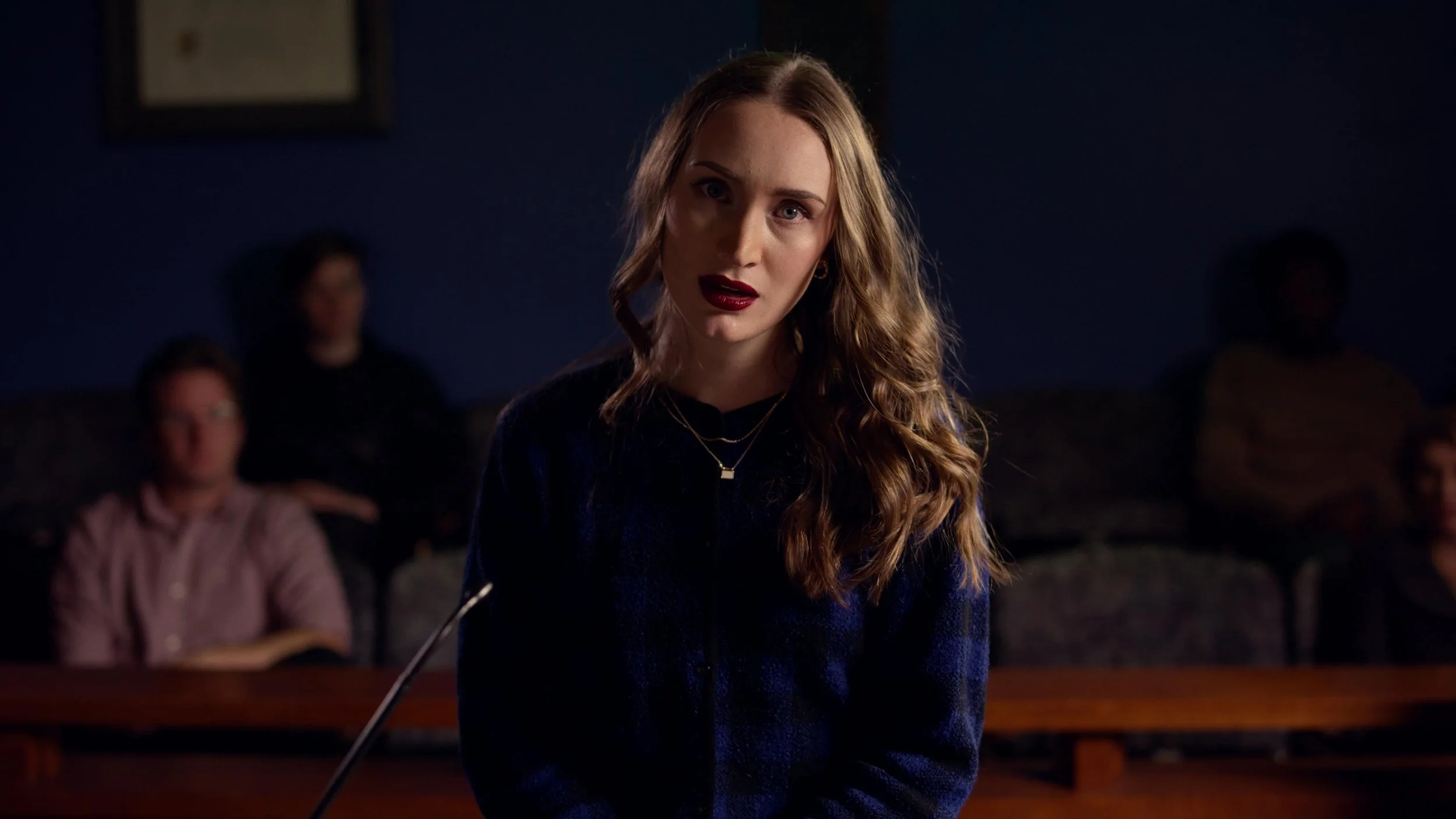 A young woman with wavy brown hair and red lipstick, sitting in a dark room with blurred people in the background.