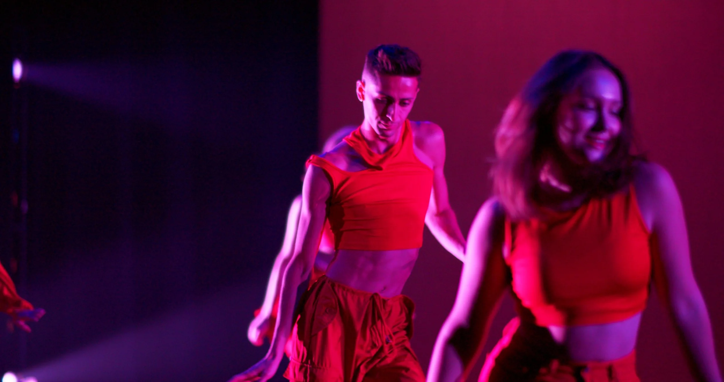 Two dancers in red outfits performing on stage under purple and pink lighting, with one dancer in the foreground smiling and a male dancer behind her