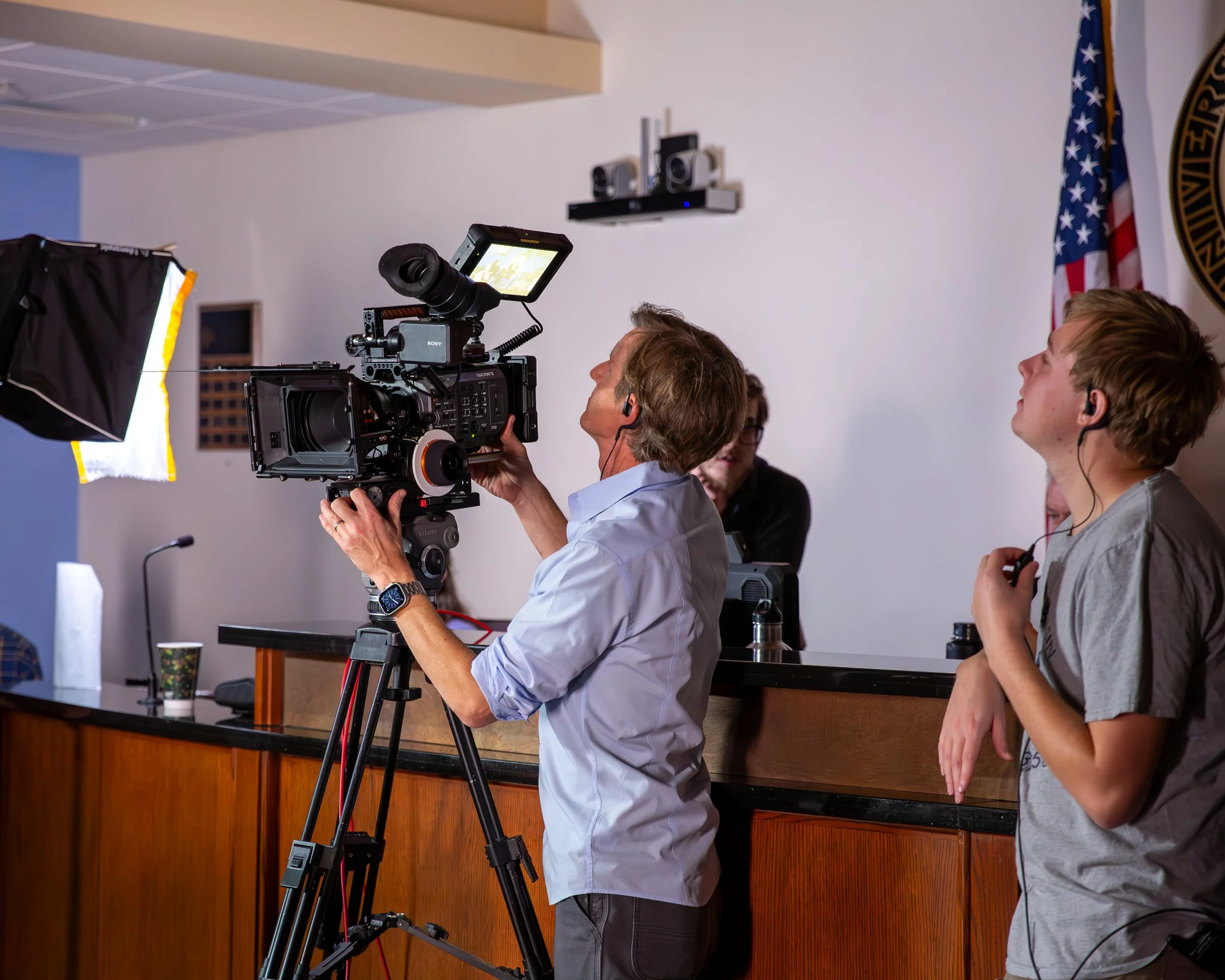 Filmmaker with professional camera filming at a press event or conference with individuals wearing headphones, in a room with an American flag and a seal on the wall.