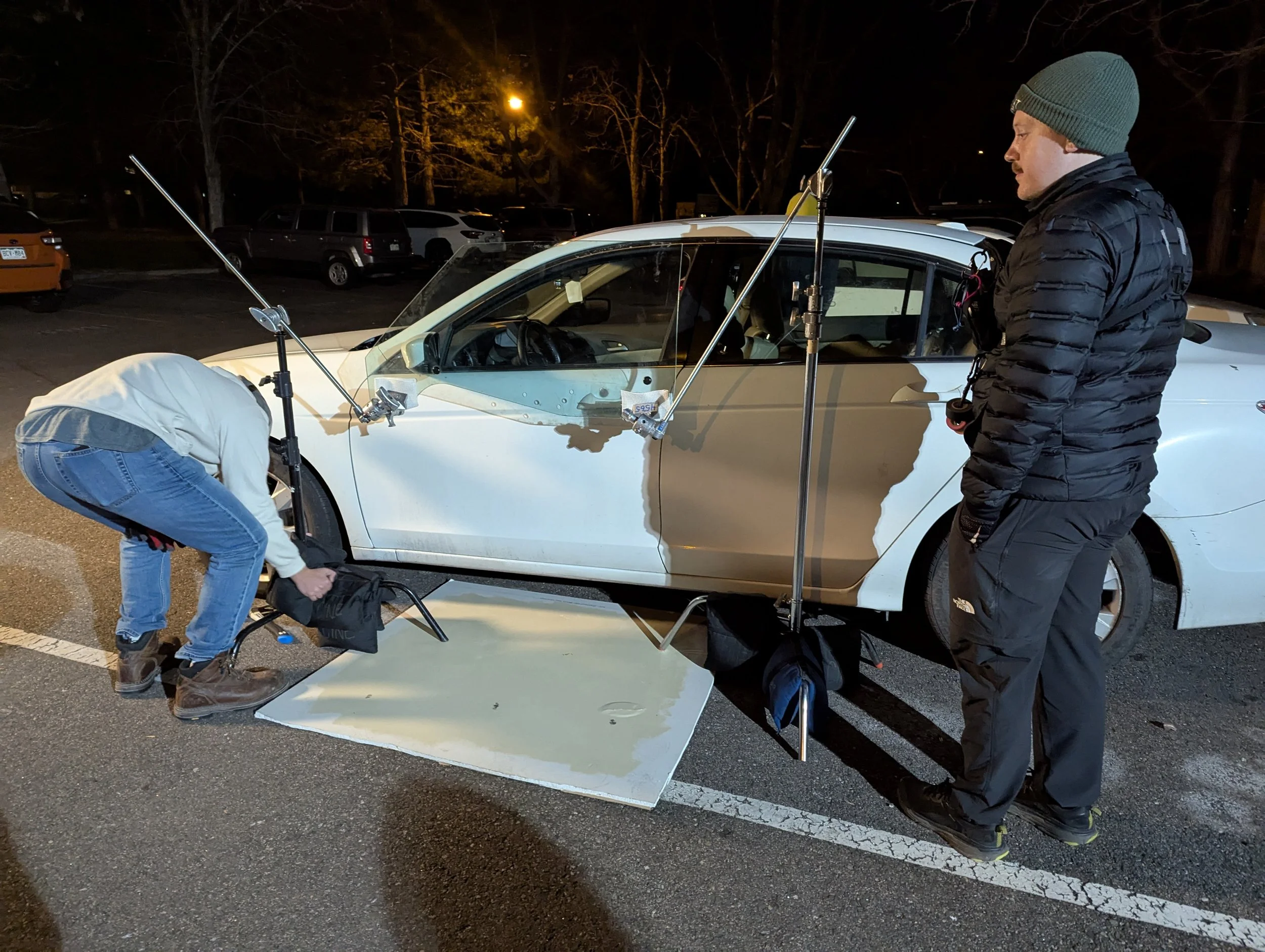 Two forensic investigators examine a white car in a parking lot at night, with one person bent over and the other standing, surrounded by equipment including a large white board and tall metal poles.
