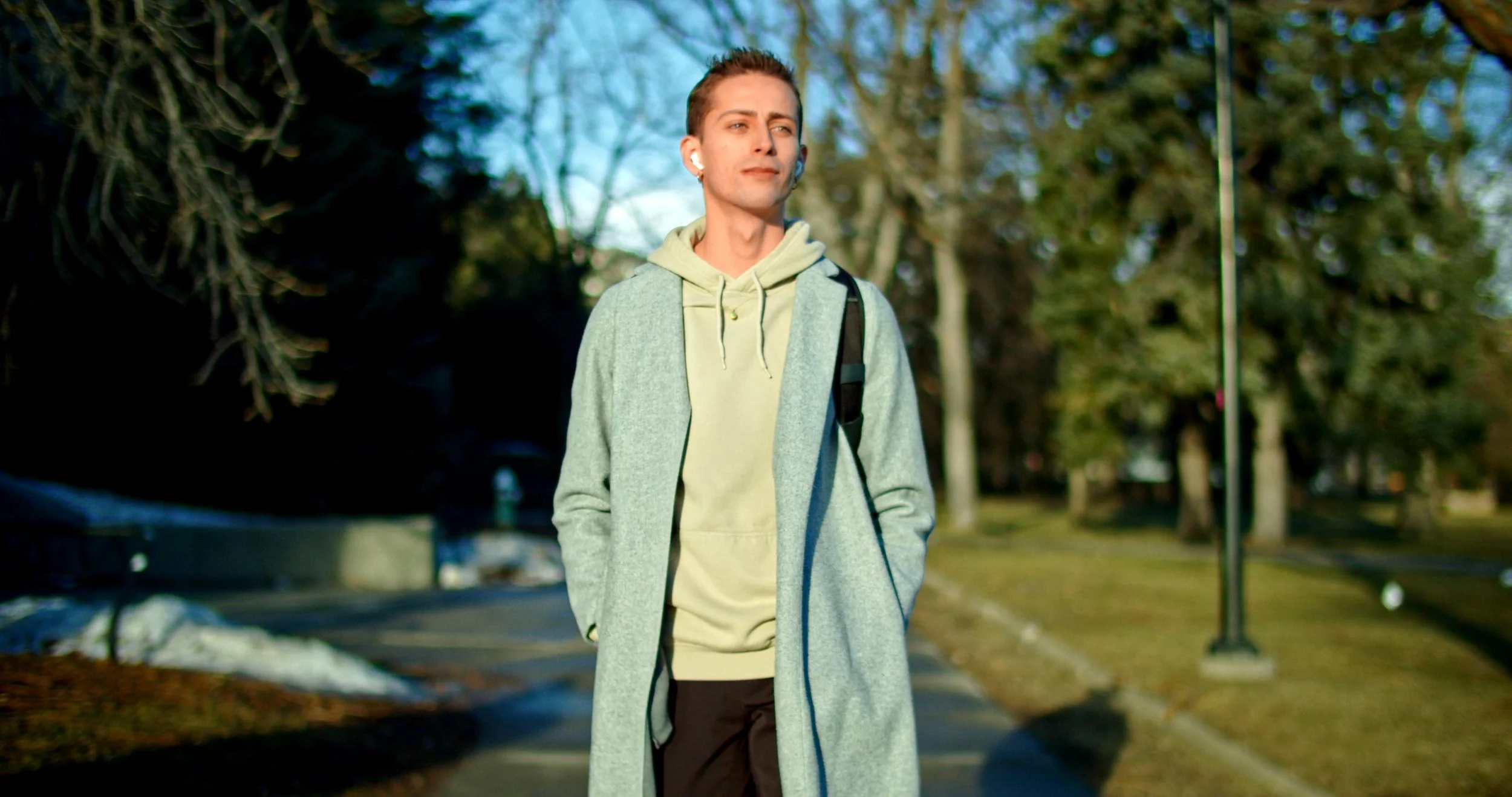 Young man walking outdoors on a sunny day, wearing a light gray coat, beige hoodie, black pants, carrying a backpack, and listening to earbuds.