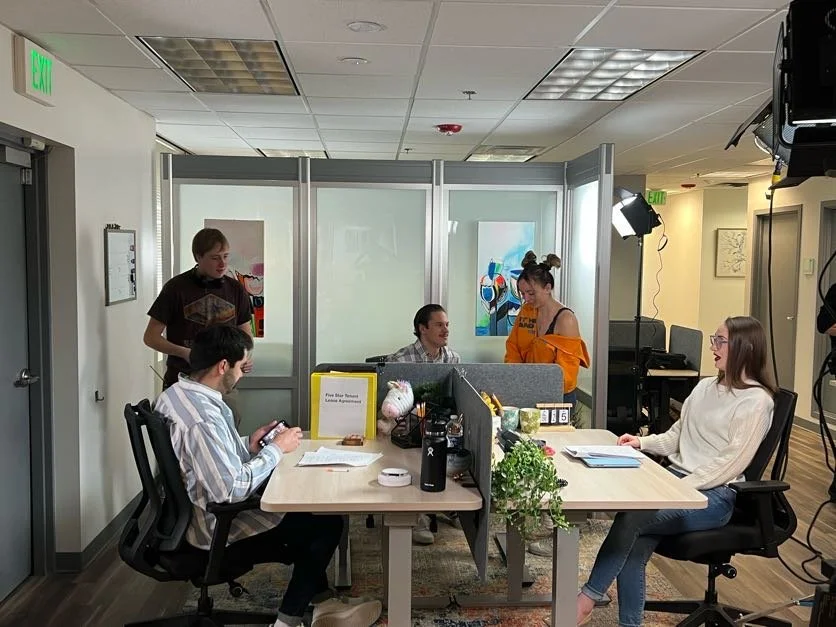 Office with five people, two sitting at a desk, and three others nearby, one standing and two behind a partition, in a modern workspace.