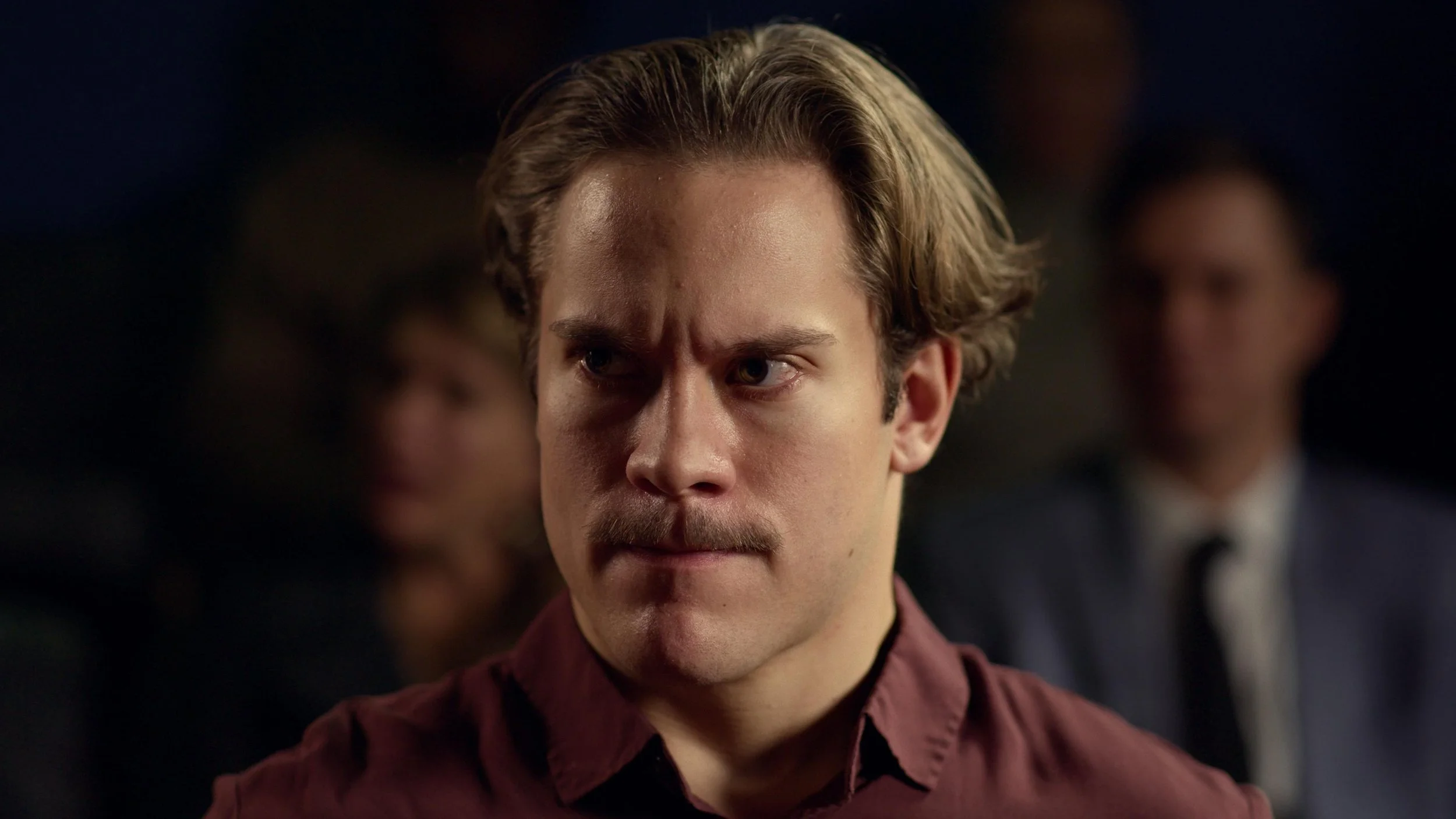 A man with light skin, brown wavy hair, and a mustache, looking serious with a focused expression, wearing a maroon shirt, in a dimly lit environment with blurred background figures.