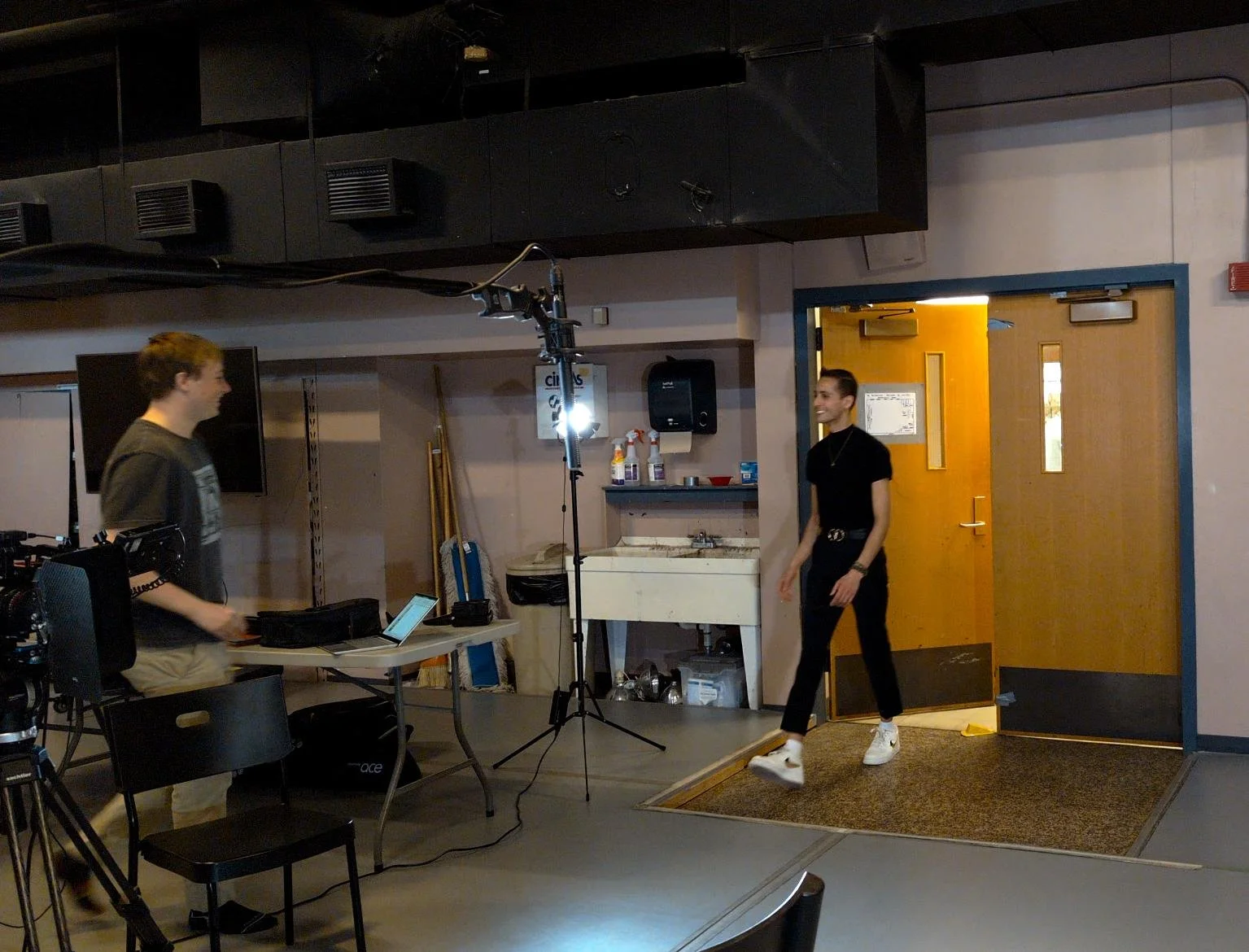 Two young men on a video set with film equipment; one is walking out of a door into a room, and the other is operating a camera. The scene appears to be part of a film or video shoot.