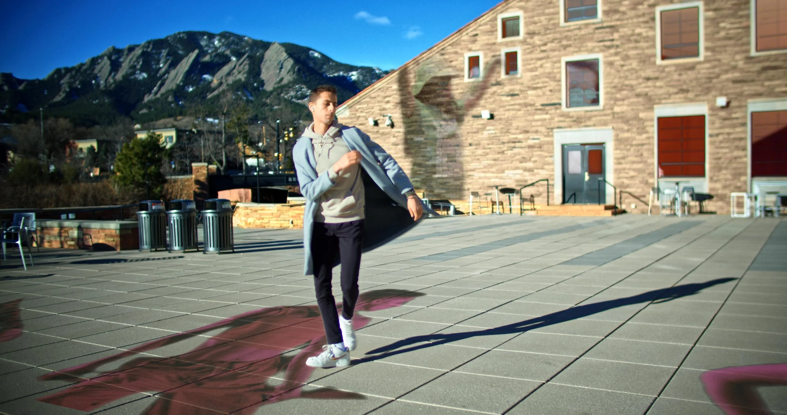 A young man dressed in casual athletic attire walking outdoors in front of a brick building with mountains in the background.
