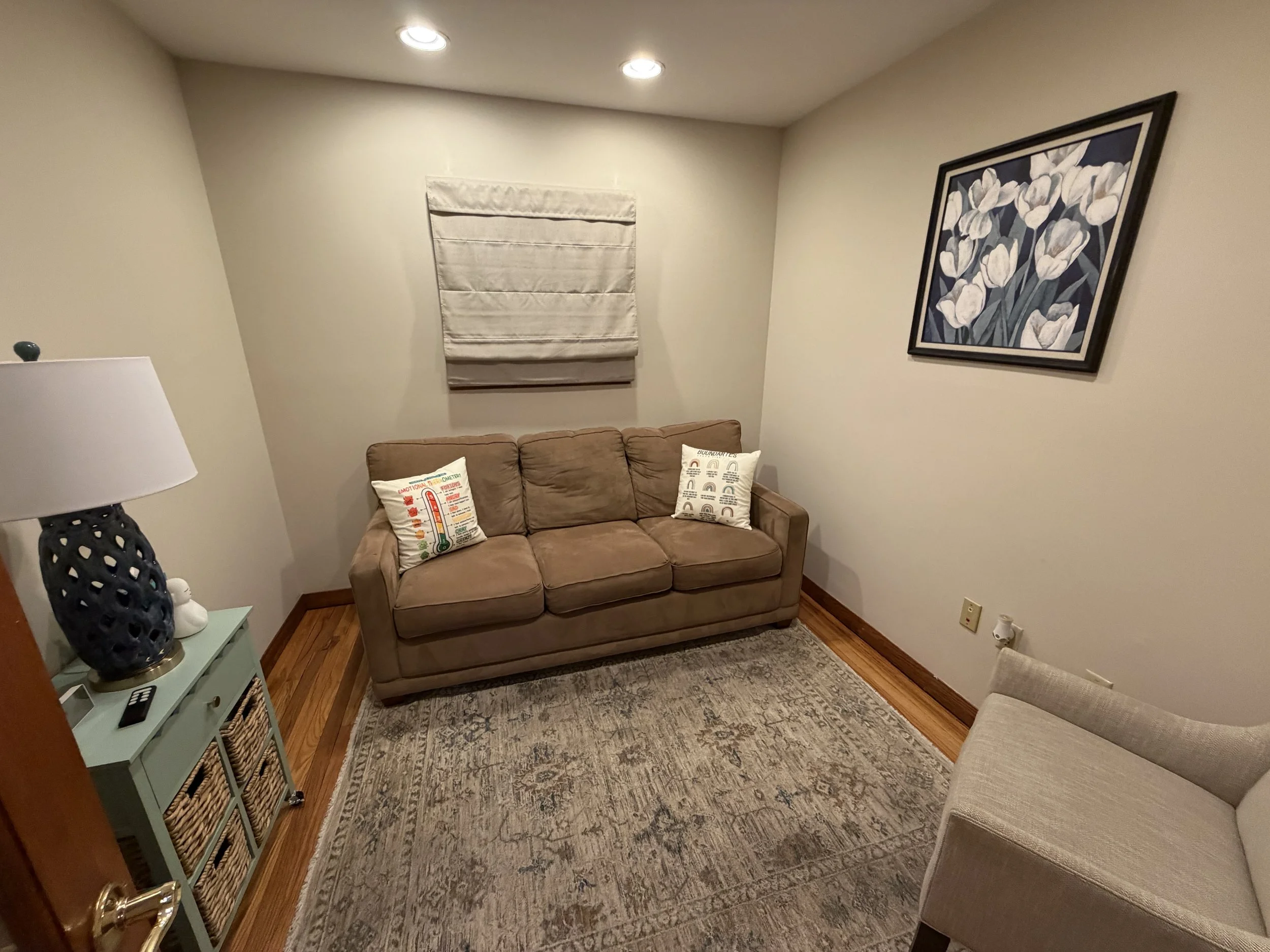A cozy living room corner with a beige loveseat sofa, decorated with two embroidered pillows. A framed floral painting of white tulips hangs on the wall above. To the left, a side table holds a modern black lamp with a white lampshade and a decorativ