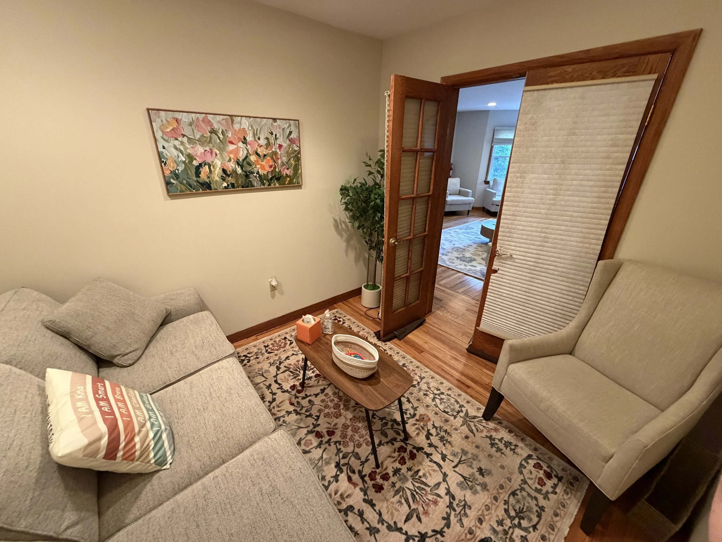 Living room with a beige sofa, a pillow with rainbow stripes, a wooden coffee table with a tissue box and basket, a beige armchair, a patterned rug, a wall art depicting pink and orange flowers, a potted plant, a partially opened wooden door, and ano