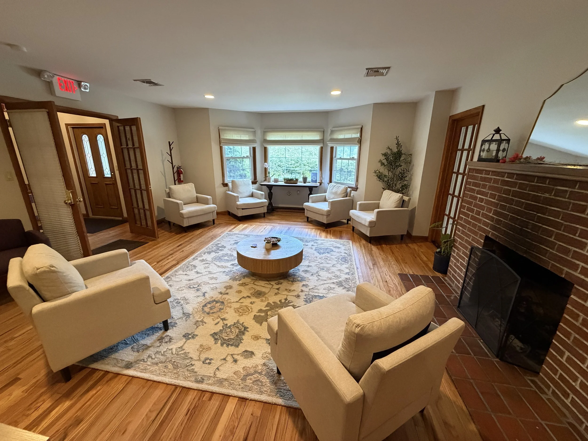 Living room with beige armchairs, a patterned area rug, a brick fireplace, and large windows with white blinds, decorated for the holidays.