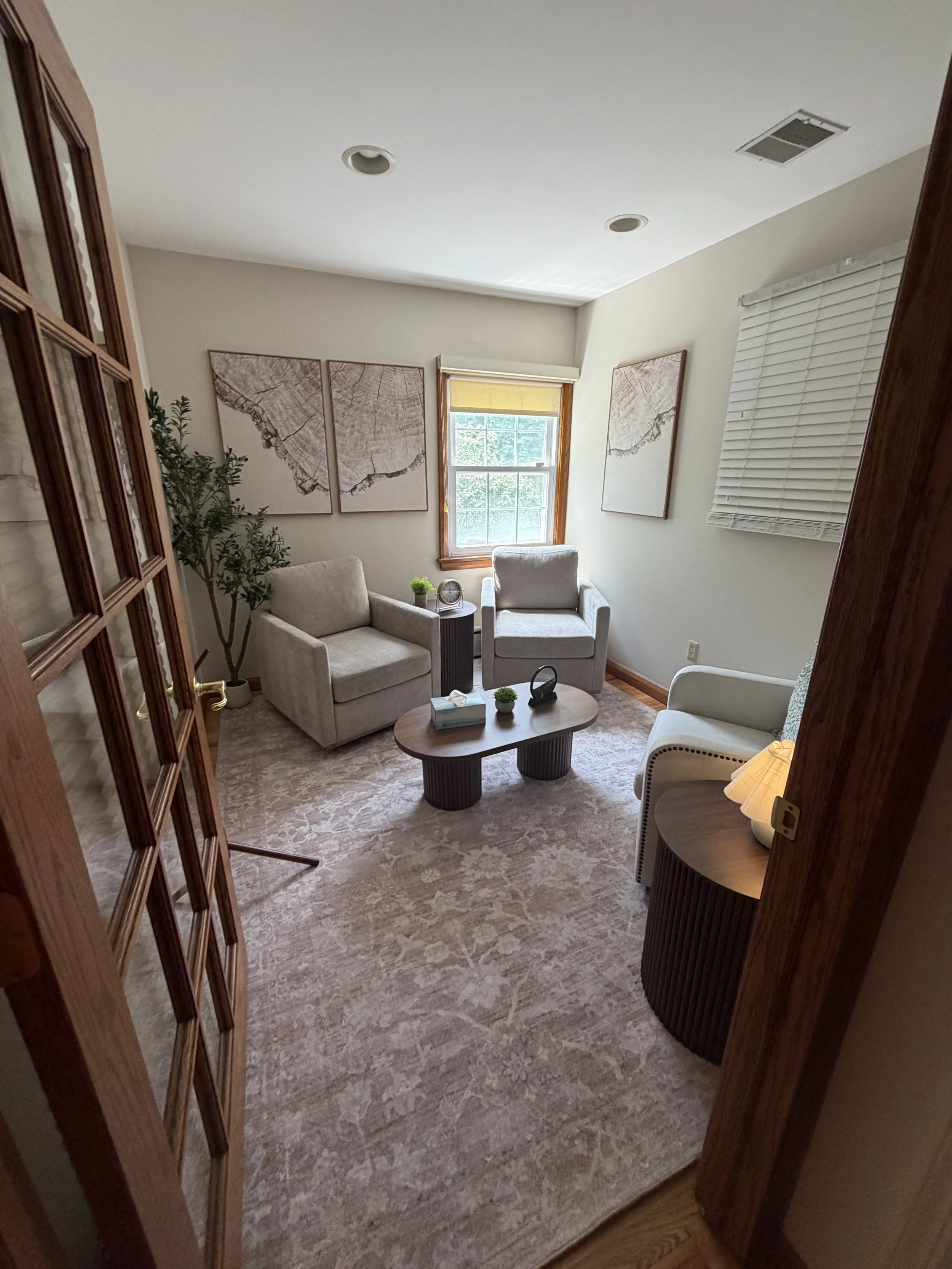 A cozy sitting room with two beige armchairs and a small round coffee table, decorated with plants and artwork, with a window and blinds, and a patterned rug.