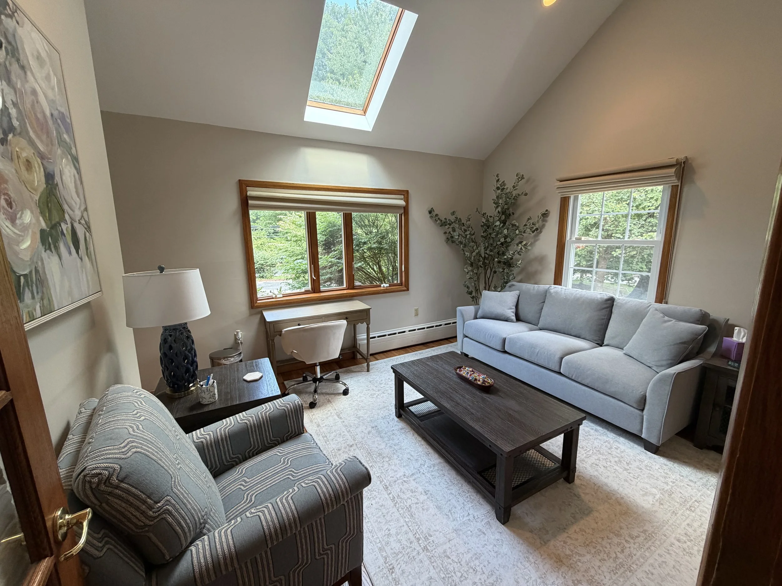 A living room with a high, angled ceiling and a skylight. There are two windows with shades, a gray sofa with pillows, a patterned armchair, a black coffee table, a desk with a chair, a table lamp, a large painting on the wall, and decorative greener