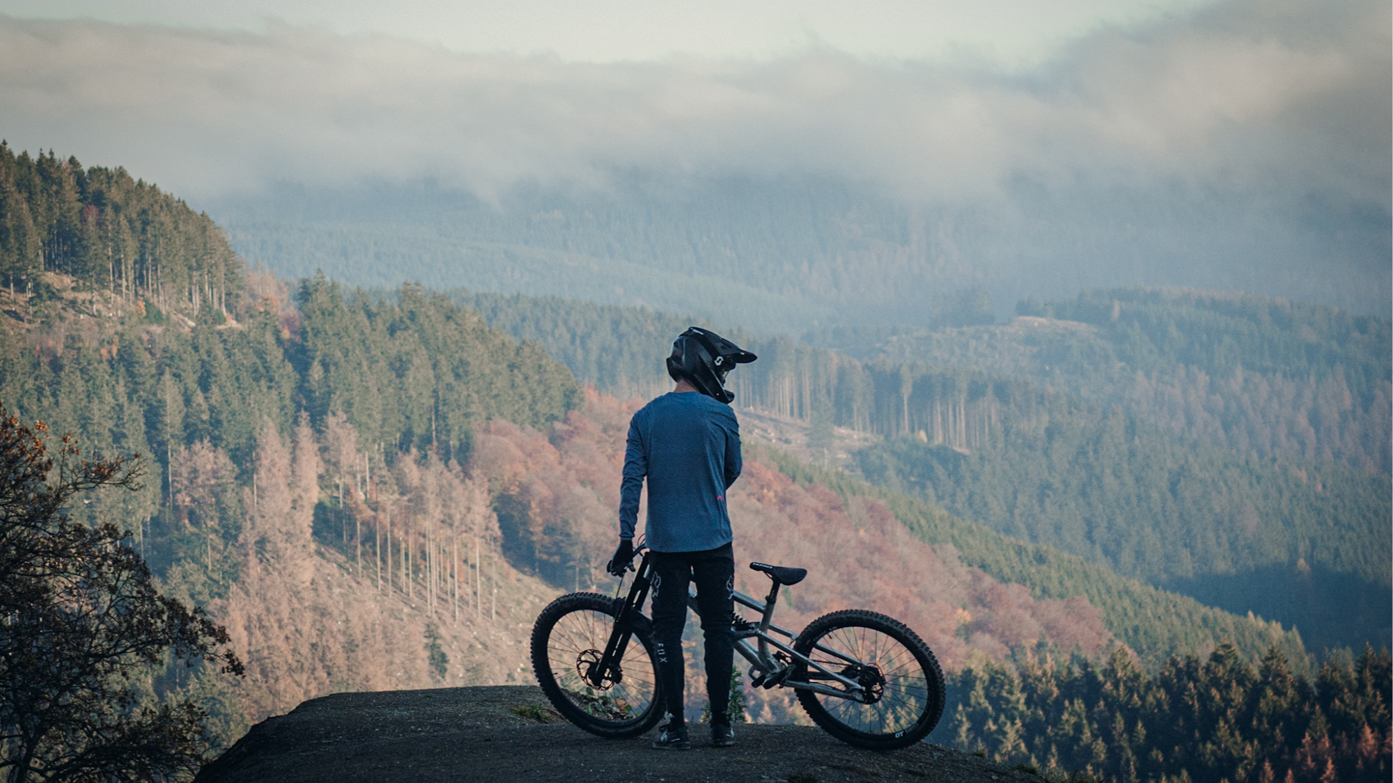 Ein Mountainbiker steht mit seinem Bike auf einem Felsen und blickt auf eine bewaldete Berglandschaft mit Wolken am Himmel.