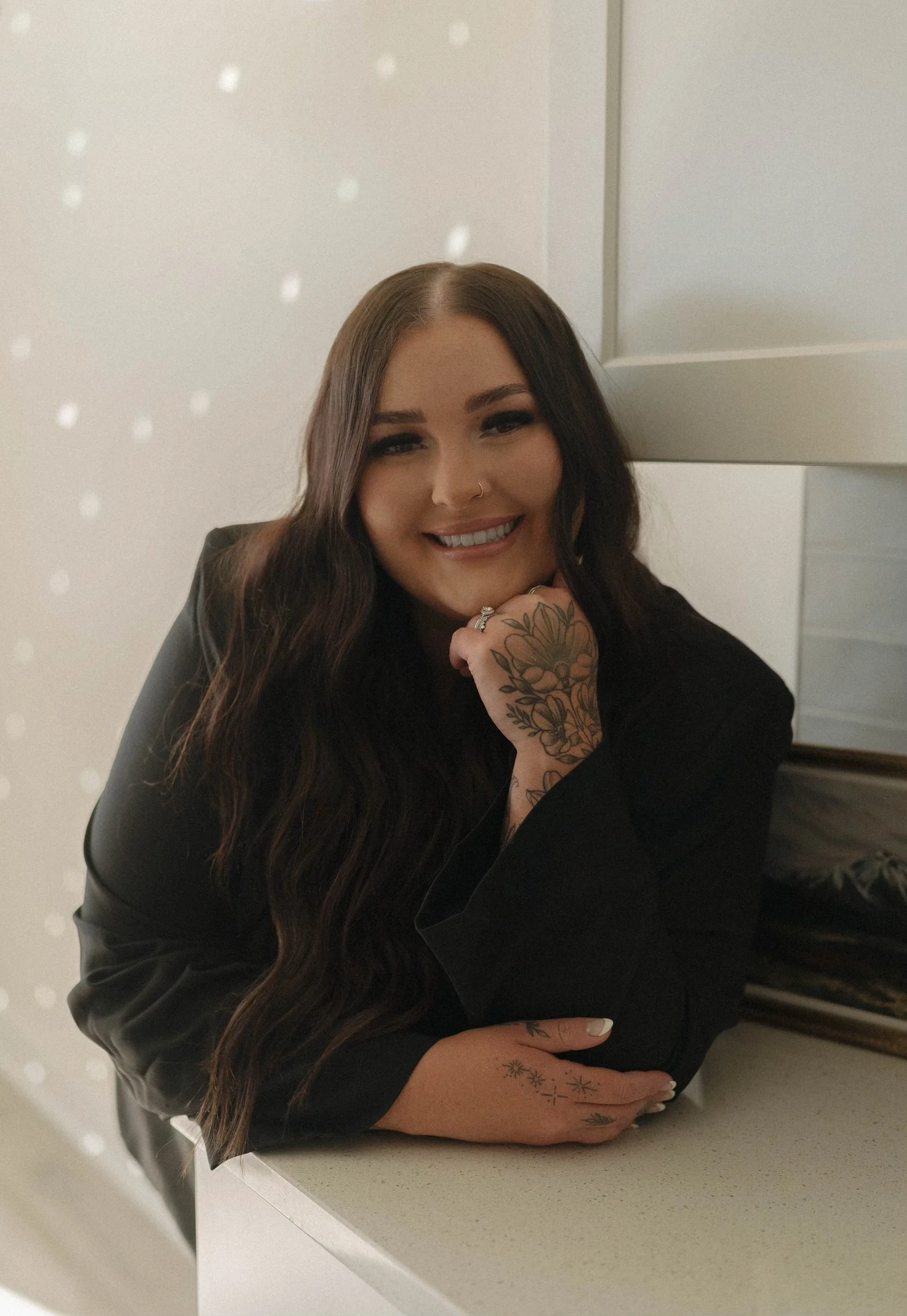 A woman with long, dark hair smiling, wearing a black top, with tattoos on her hand and wrist, sitting at a white countertop with a mirror and white wall in the background.