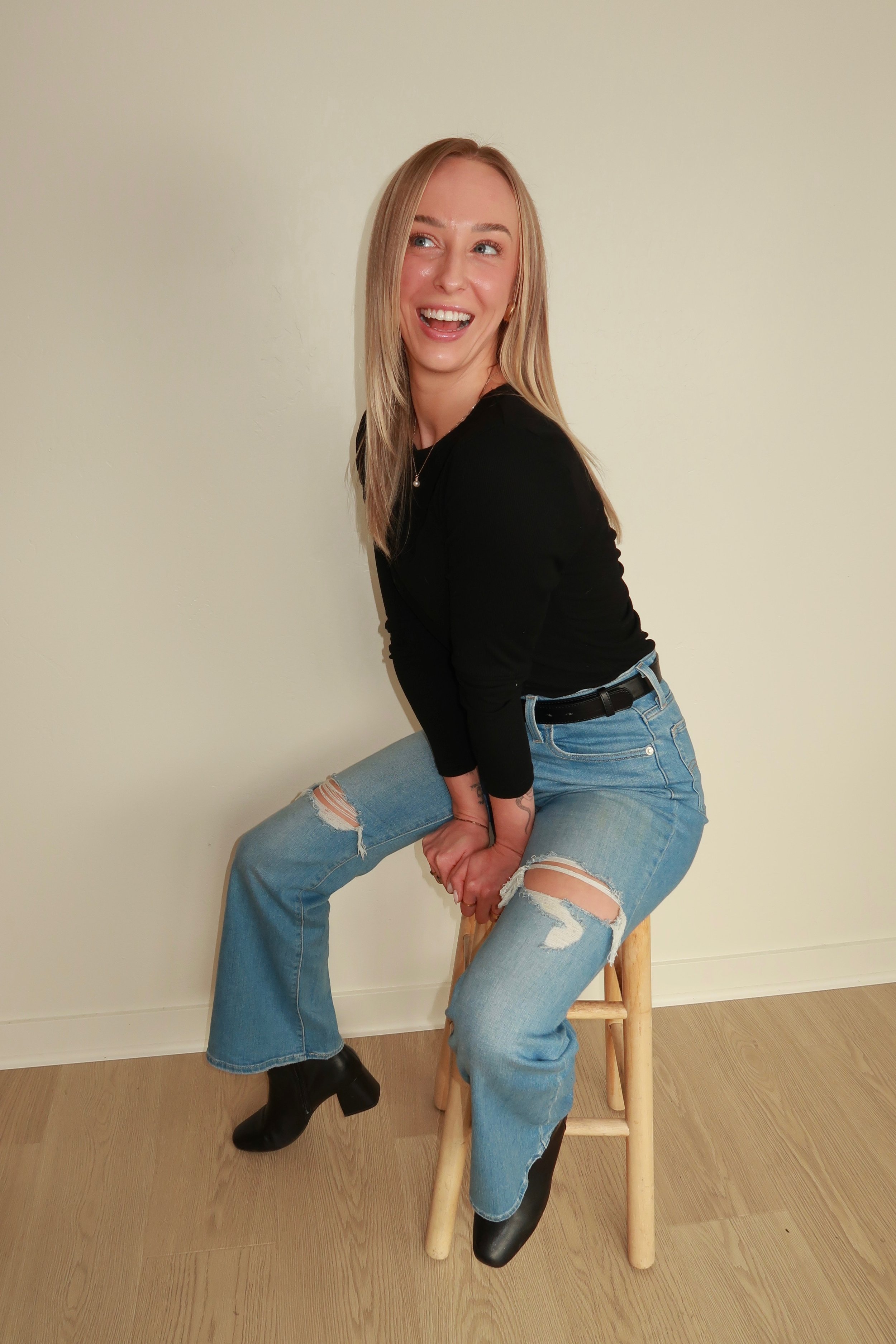 A woman with blonde hair and light skin wearing a black top, ripped jeans, and black boots is sitting on a wooden stool, smiling and laughing with her mouth open, set against a plain beige wall.