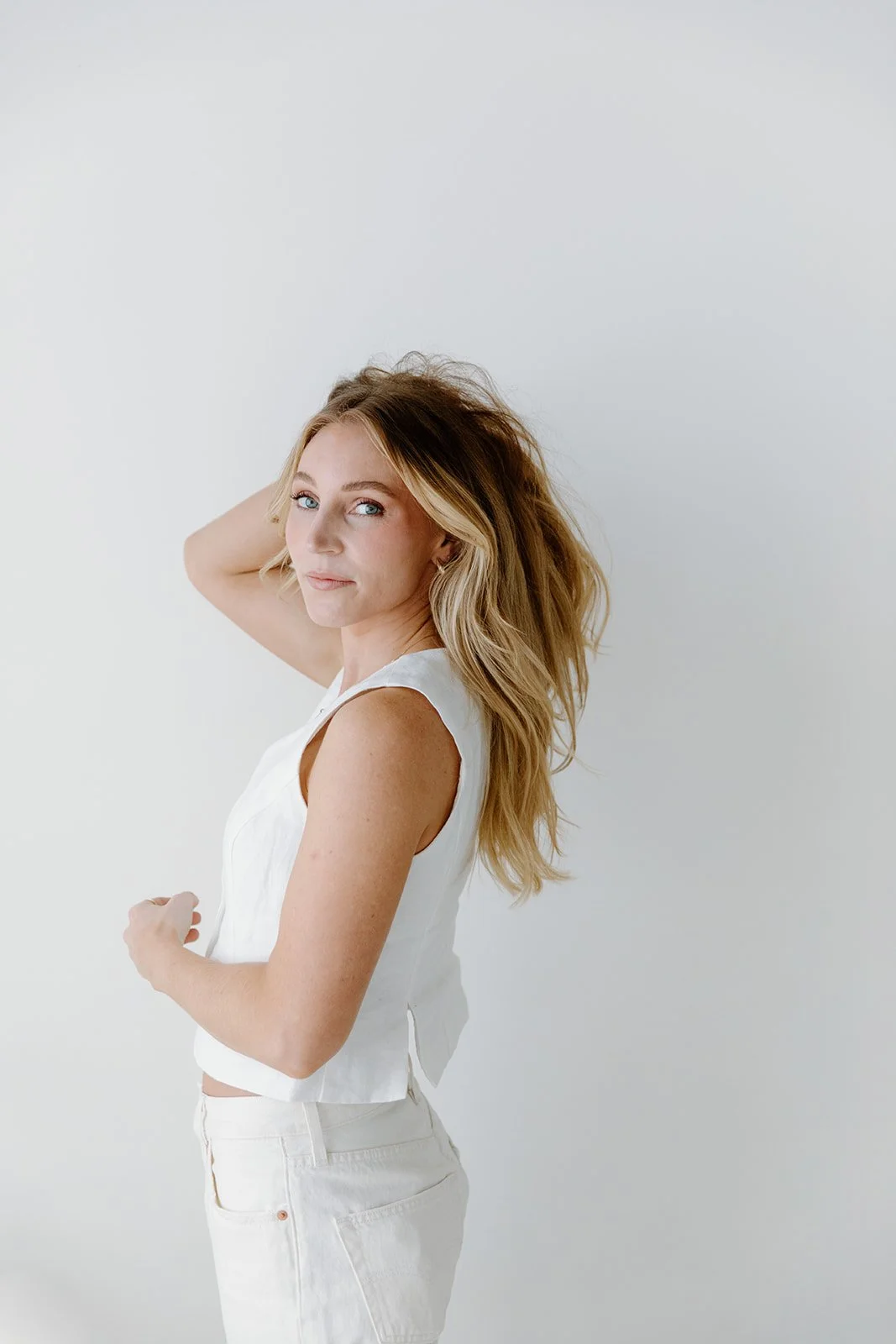 A young woman with long, wavy blonde hair looking over her shoulder, wearing a sleeveless white top and white pants, standing against a plain white background.