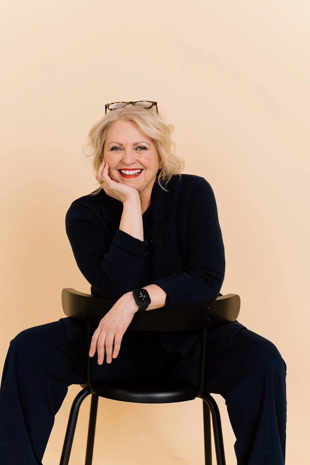 A cheerful woman with blonde hair and glasses resting on her head, sitting on a black chair against a light beige background, wearing a black outfit and a smartwatch, smiling with her chin resting on her hand.